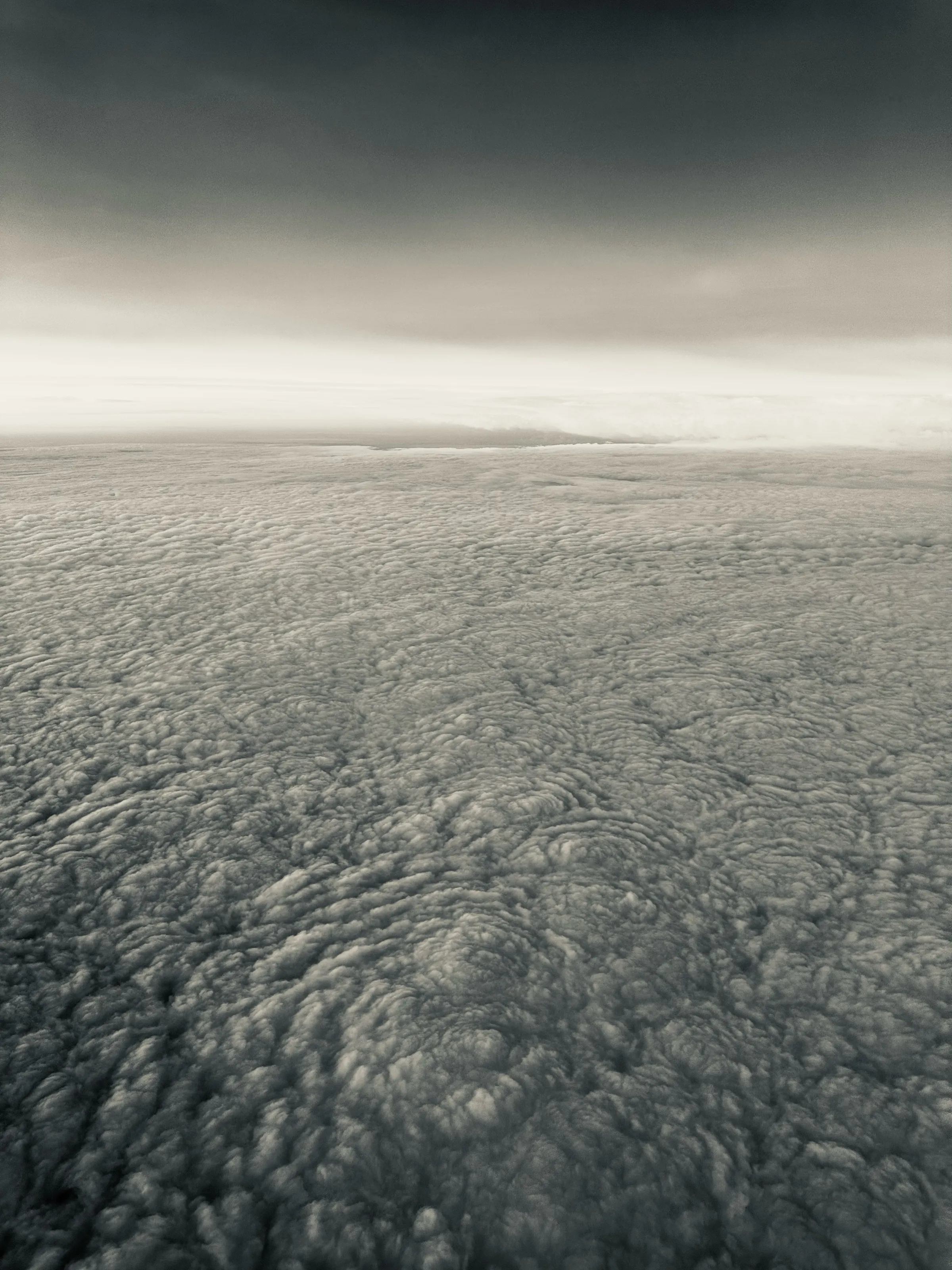 An aerial monochrome view of dense rippled clouds fading into a bright horizon under dark sky.