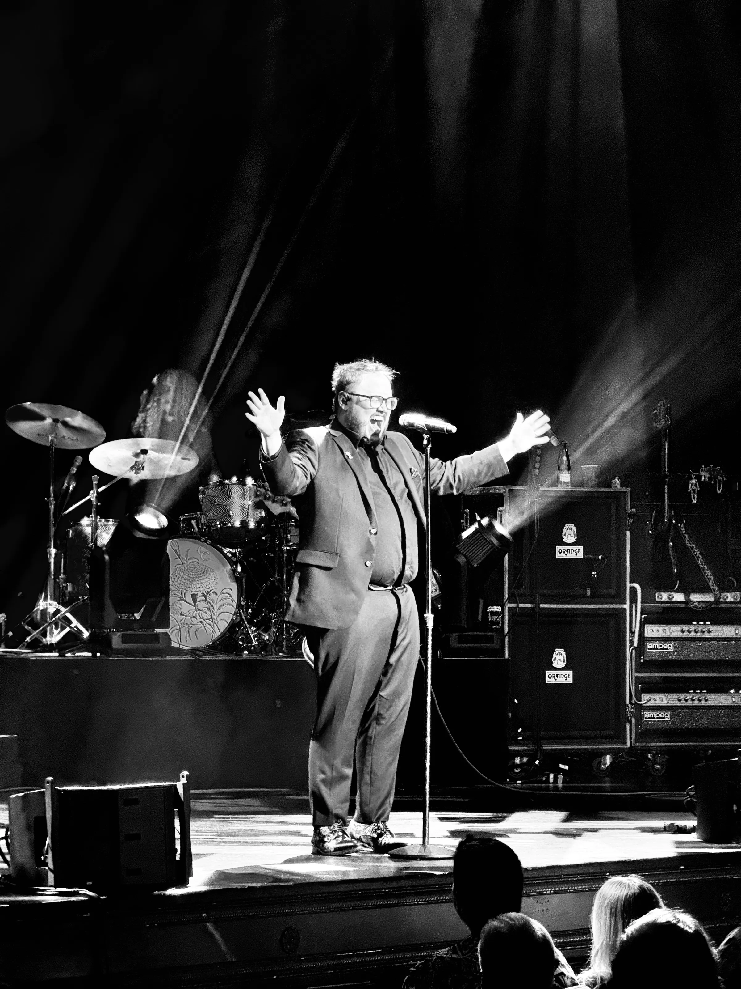 Black-and-white concert photograph of a man in a suit at a microphone with raised arms, with drums and amplifiers behind him.