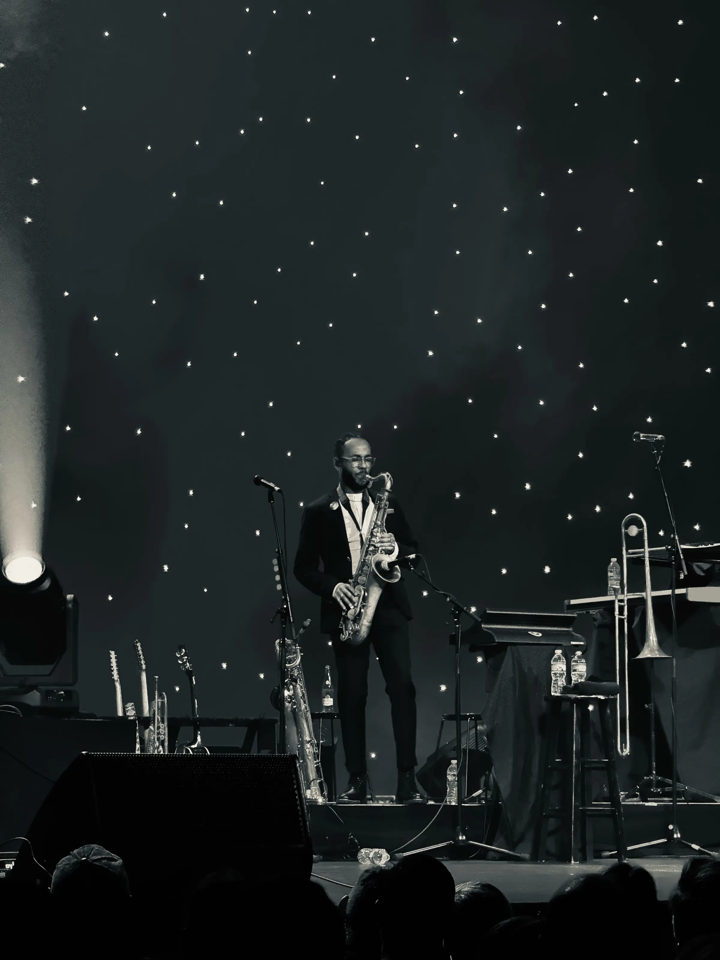 A saxophonist standing on a dark stage with instruments and small white lights behind him.