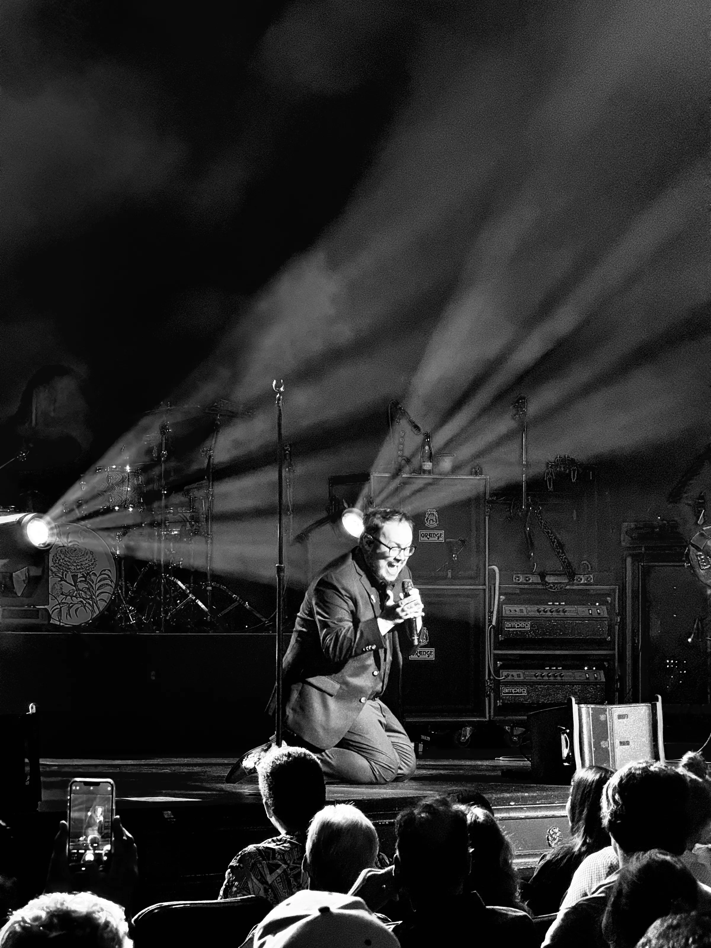 Black-and-white concert photograph of a performer kneeling with a microphone at the front of a stage, with audience heads in the foreground and diagonal beams of light behind.