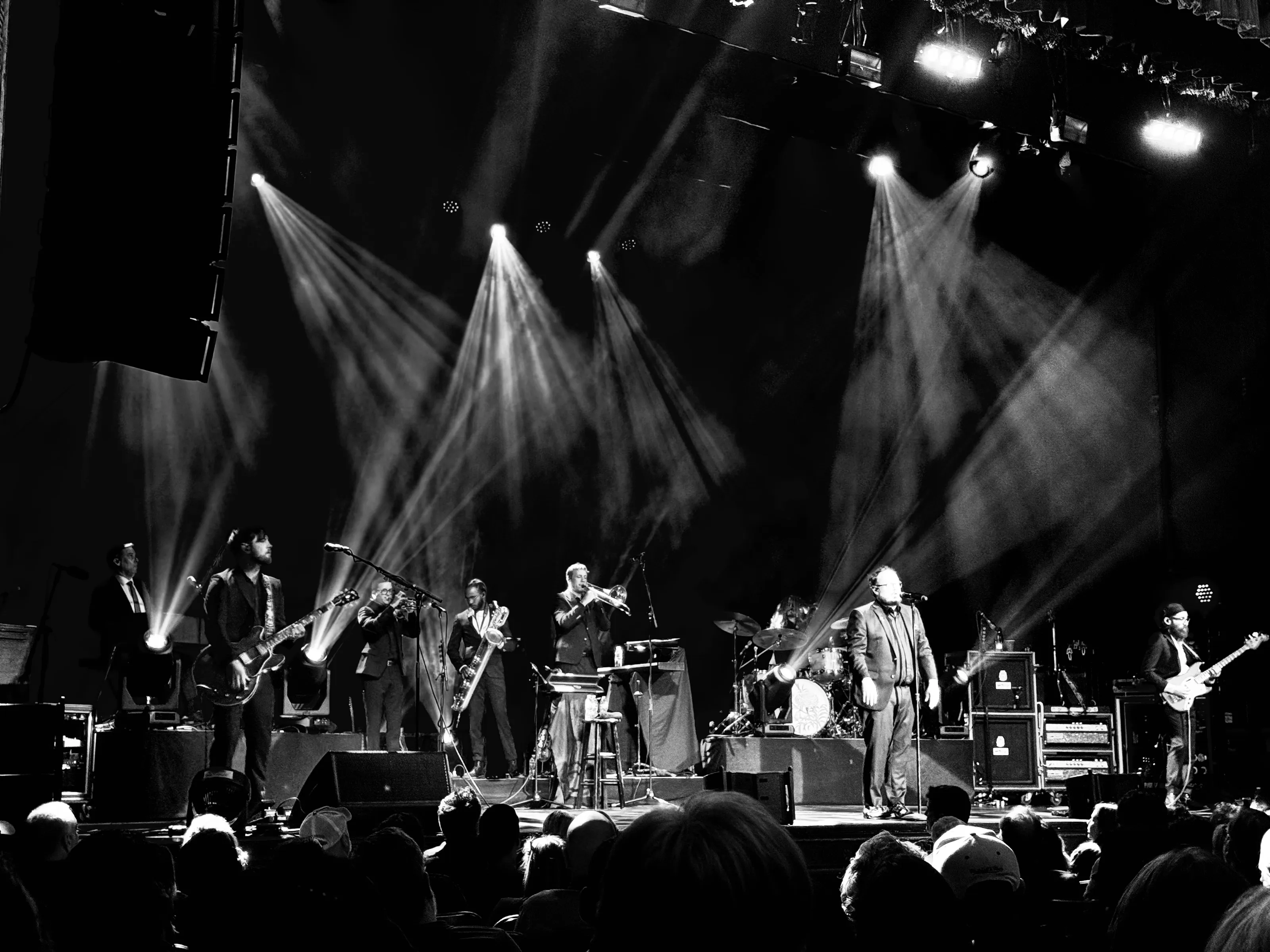 Black-and-white wide view of a band performing onstage under bright angled spotlights, with audience heads in the foreground.
