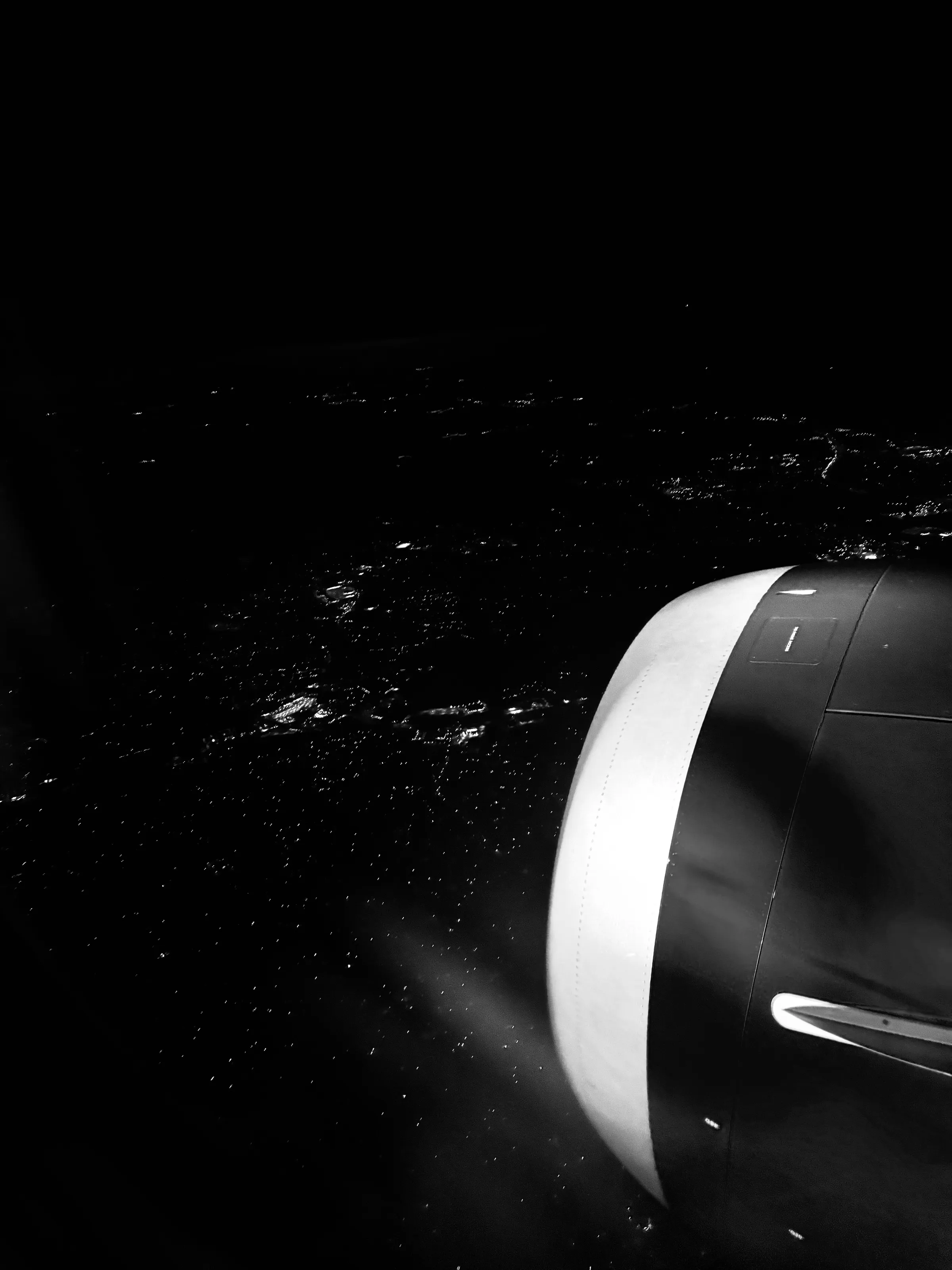 An airplane engine fills the right side of a black-and-white view of city lights at night.