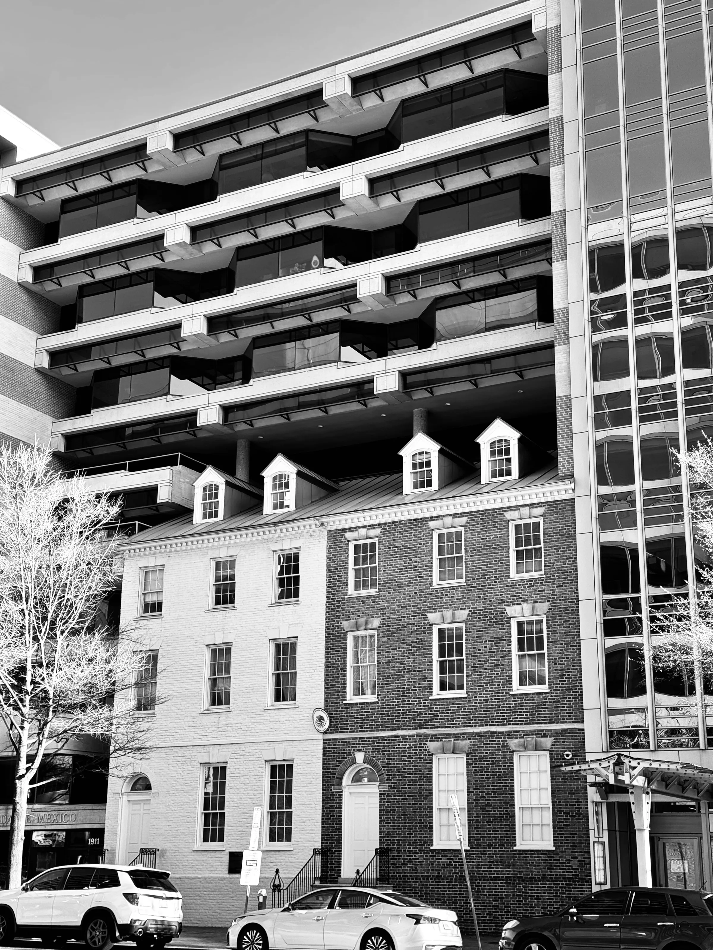 A brick row house stands beneath the stacked balconies of a much larger modern building in a black-and-white city scene.