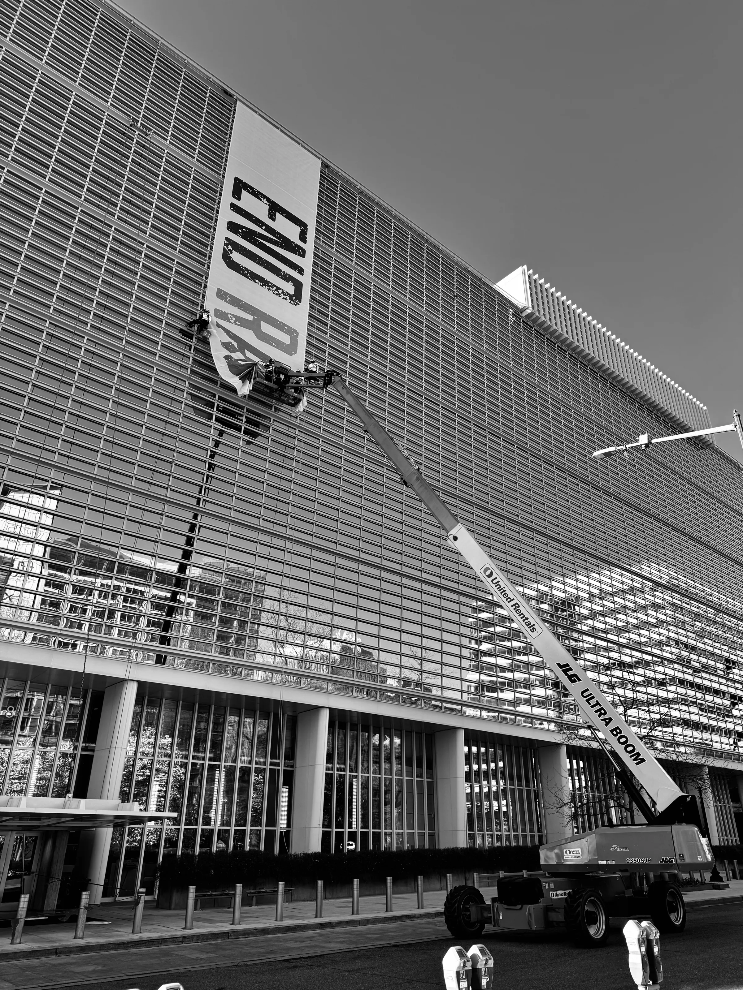 Black-and-white photograph of a boom lift raised beside a large gridded building facade and vertical sign.