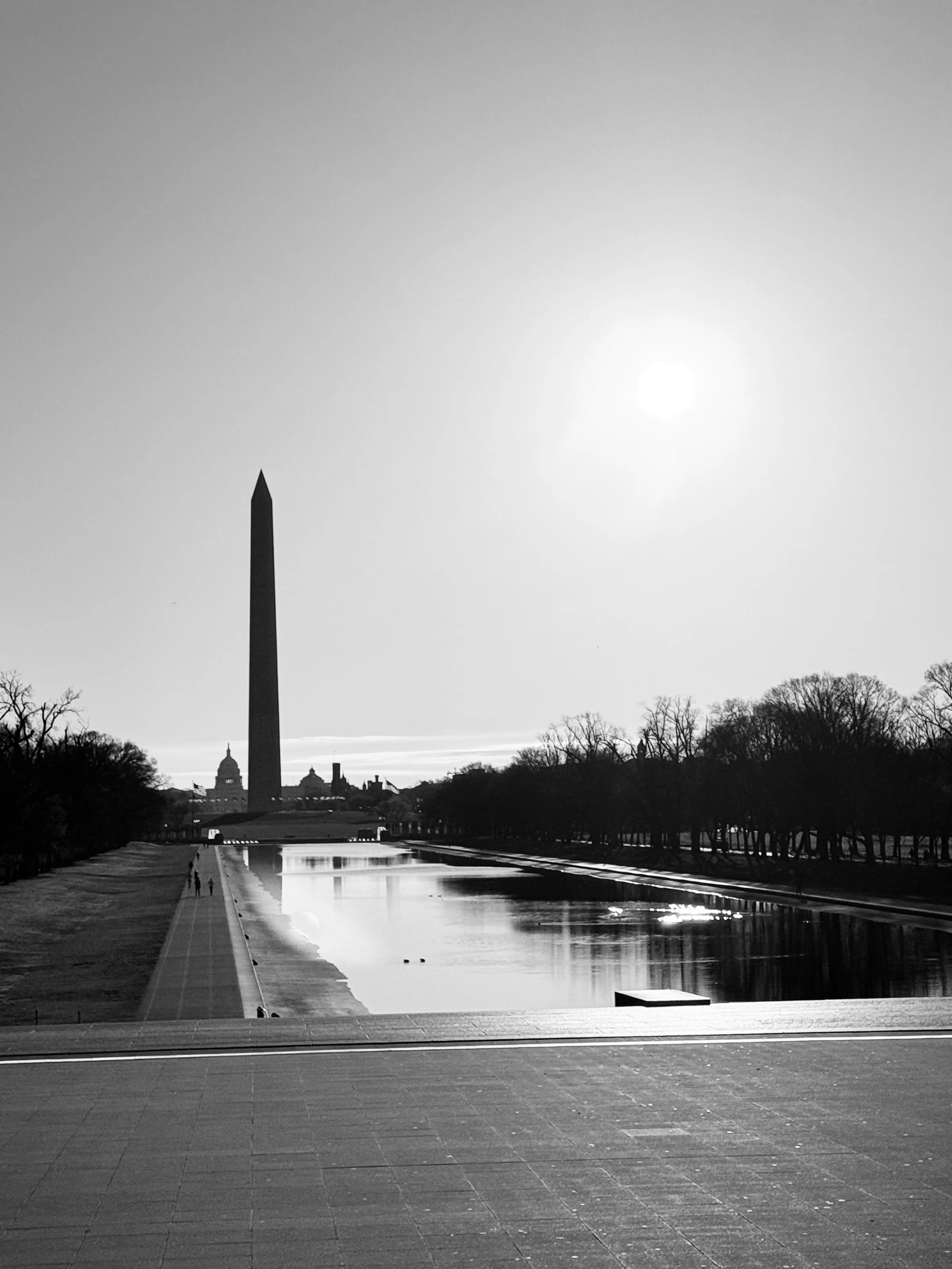 The Washington Monument rises beyond the Reflecting Pool beneath a bright sun.