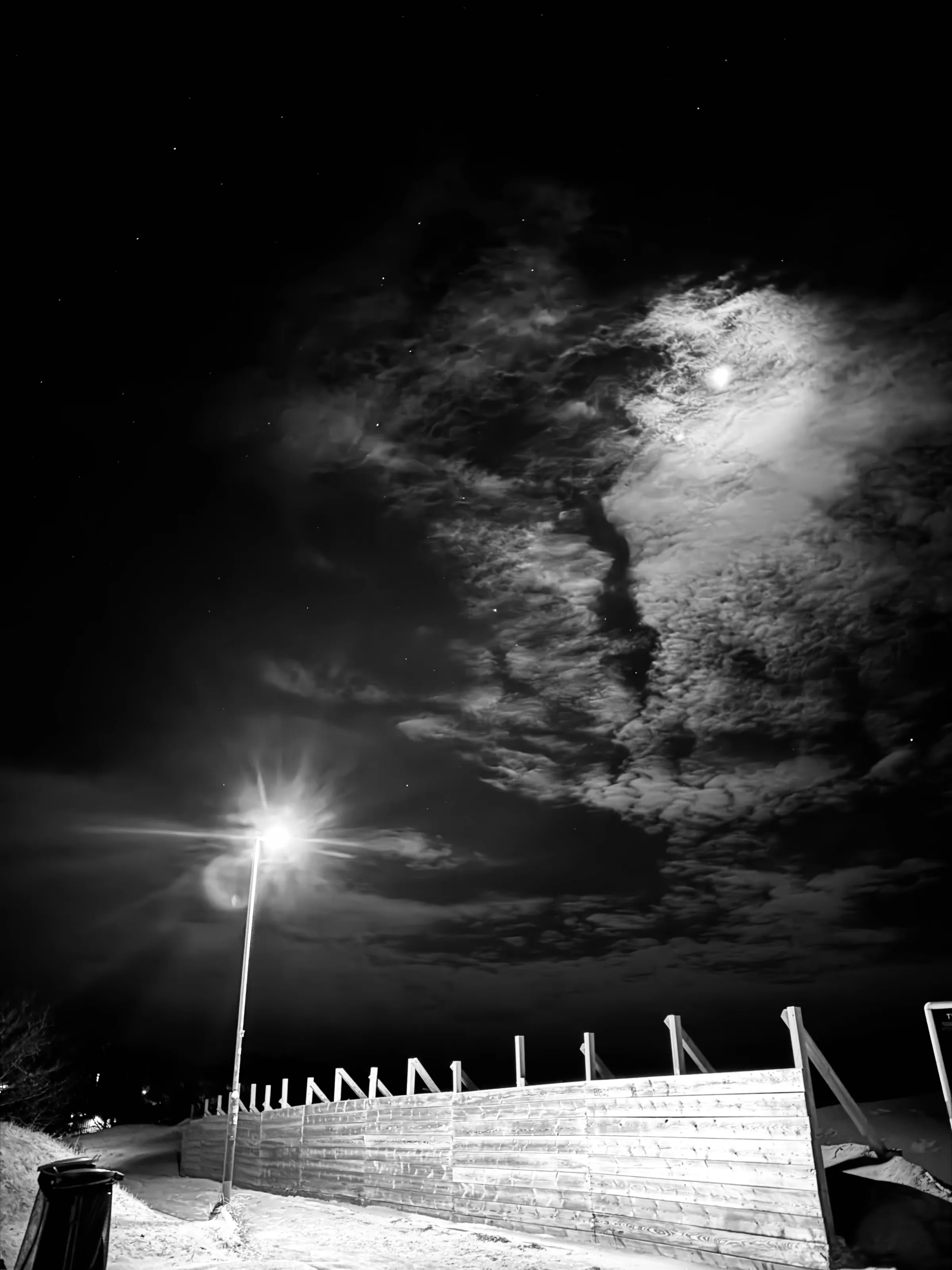 A black-and-white night scene with a bright streetlight, snow-covered barrier, and moonlit clouds.