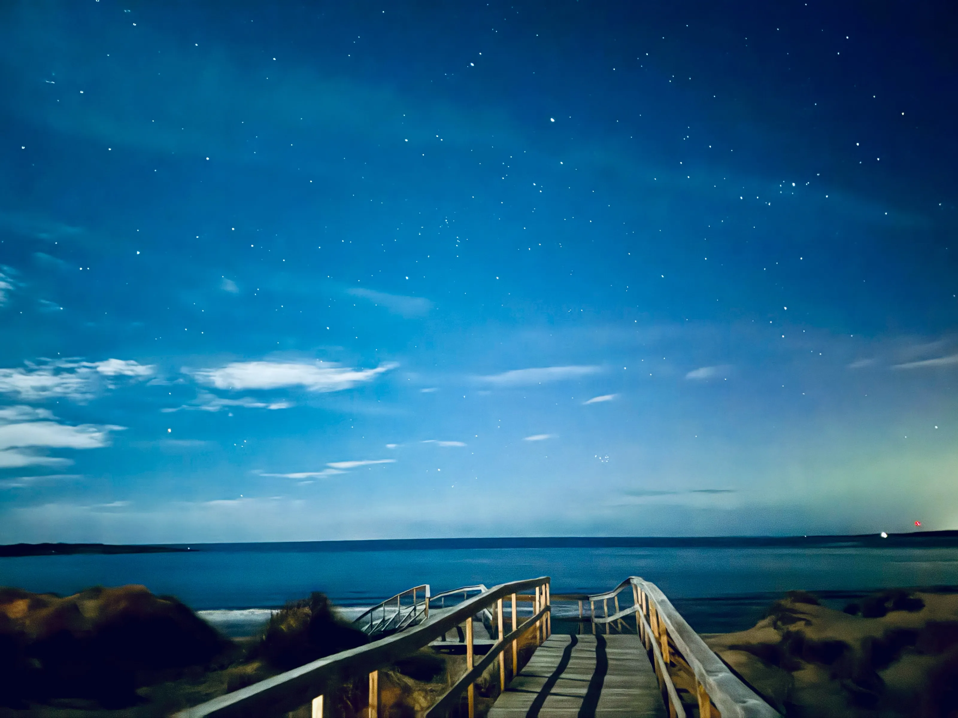 Wooden boardwalk railings lead toward a dark ocean under a blue night sky dotted with stars.
