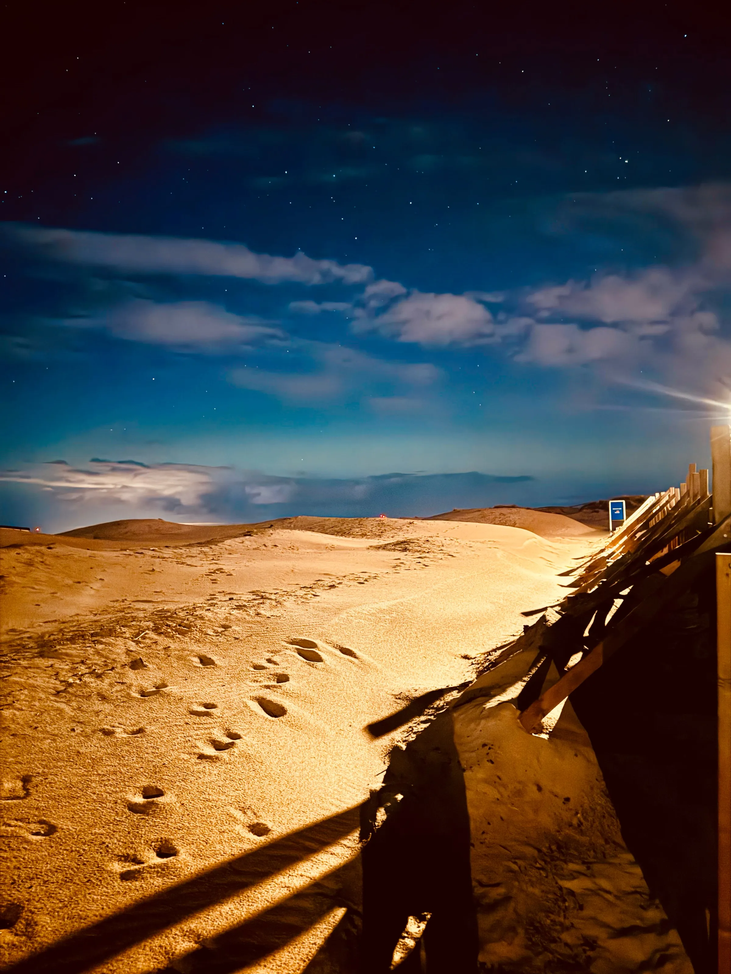 Footprints in bright sand lead toward dunes under a dark blue night sky, with a wooden walkway at right.