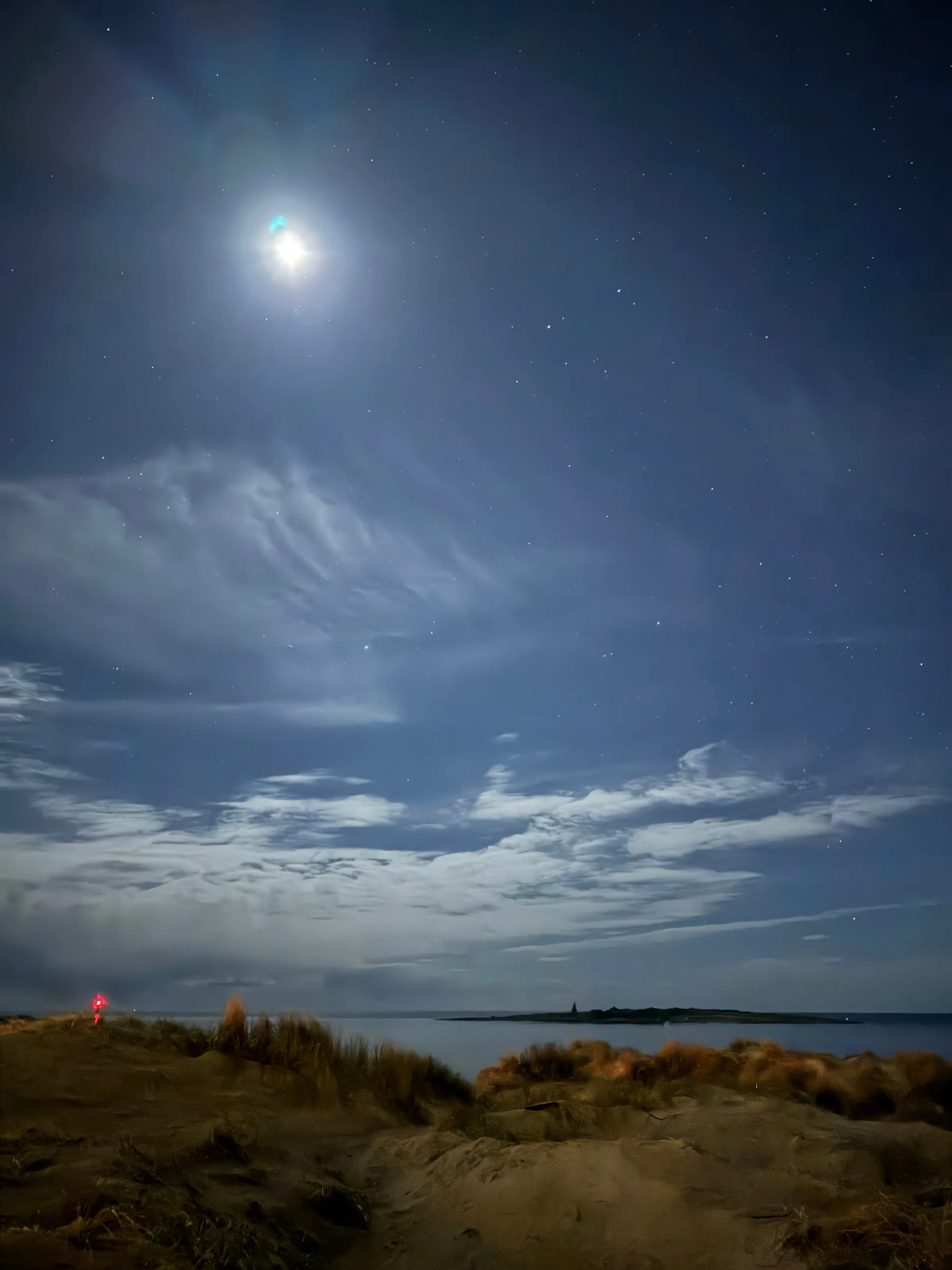 A bright moon above wispy clouds, dunes, and a distant body of water at night.