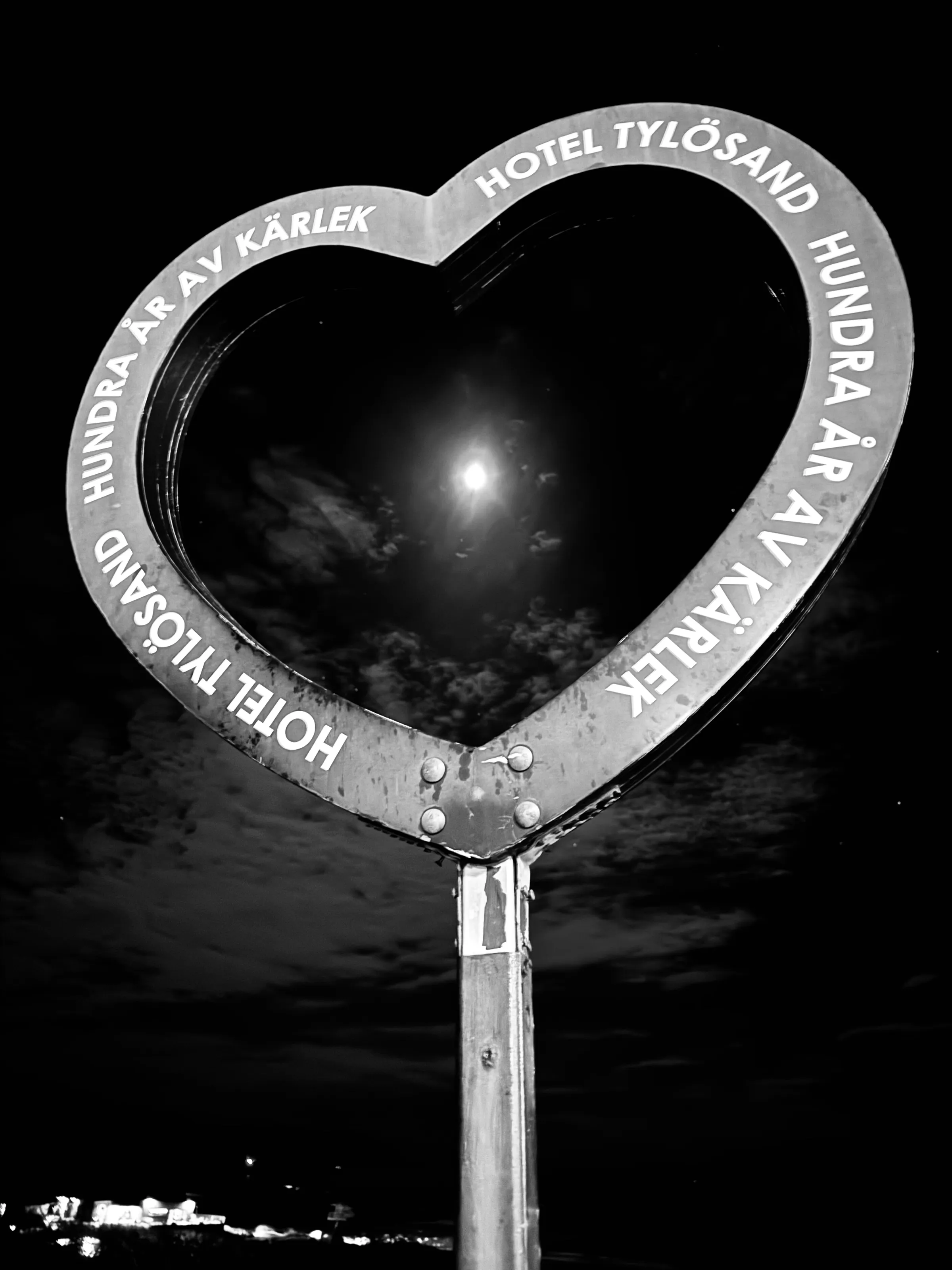 A heart-shaped metal sign with Swedish text frames a bright moon and clouds against a black night sky.