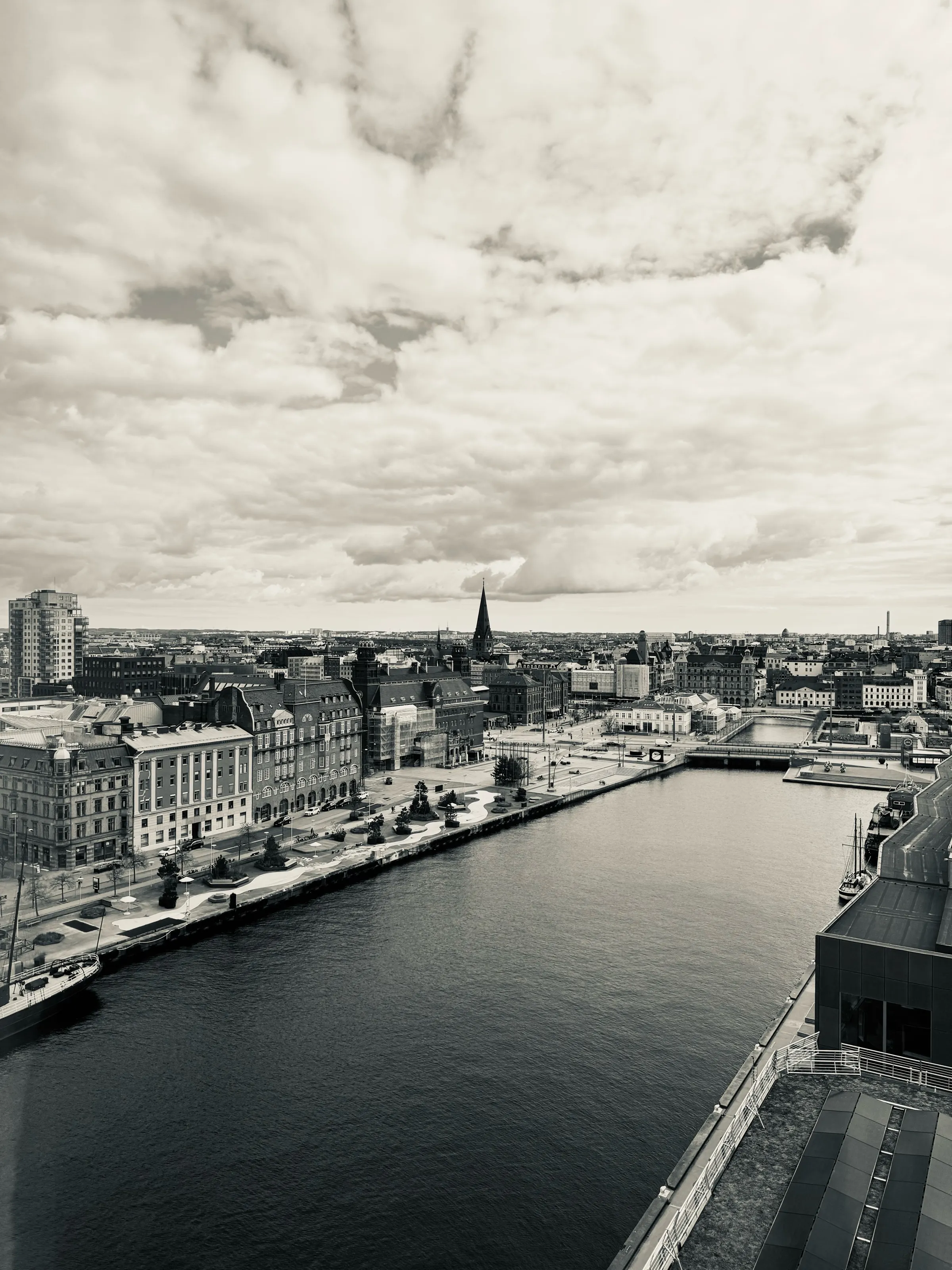 A broad canal runs through a city of low buildings and church spires beneath textured clouds.