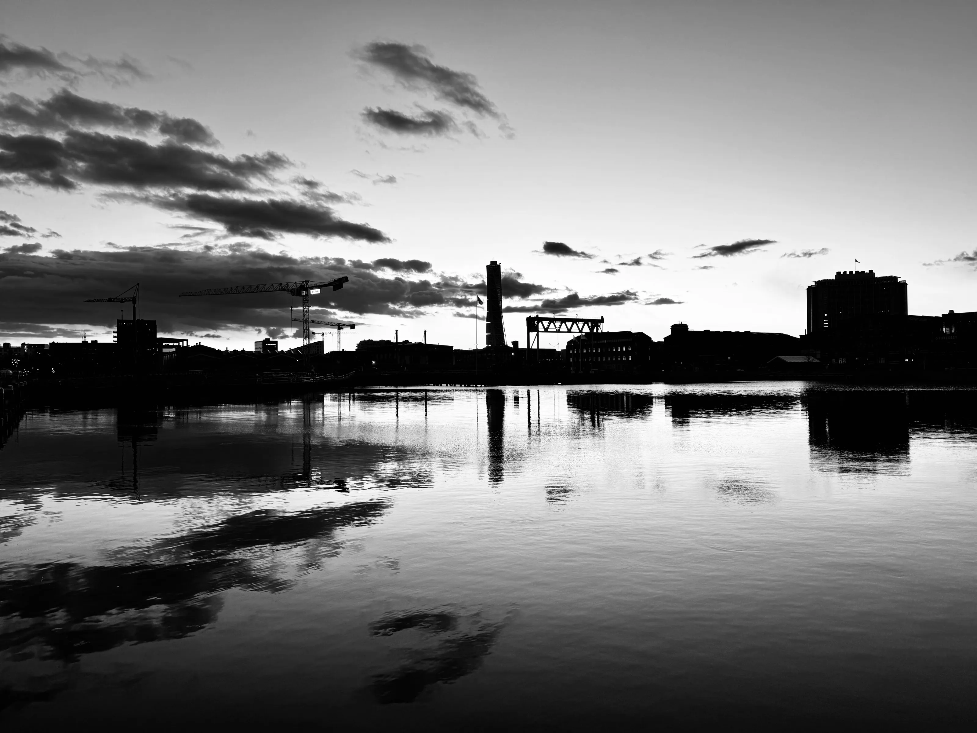 Black-and-white dusk view of cranes, low industrial buildings, and a tall smokestack reflected in calm river water.