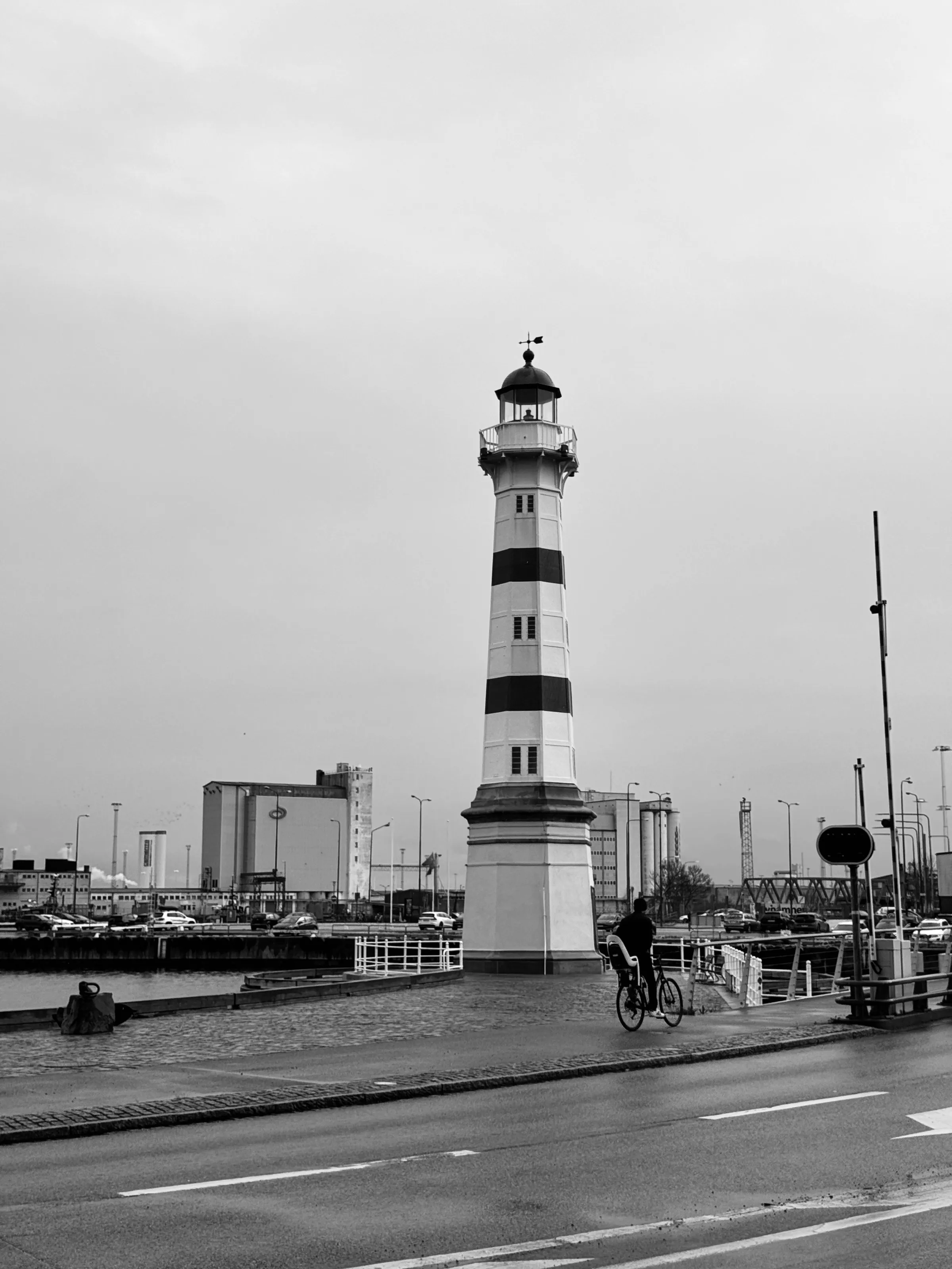 A cyclist rides past a striped lighthouse beside a harbor with industrial buildings behind it.