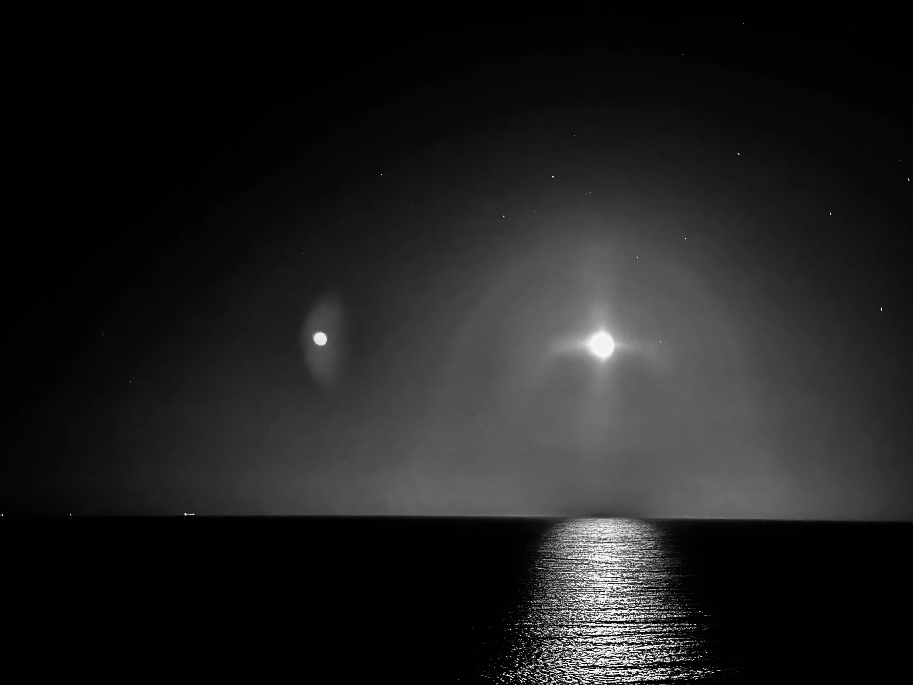 Black-and-white night seascape with two bright lights above the horizon and moonlight reflected on the water.