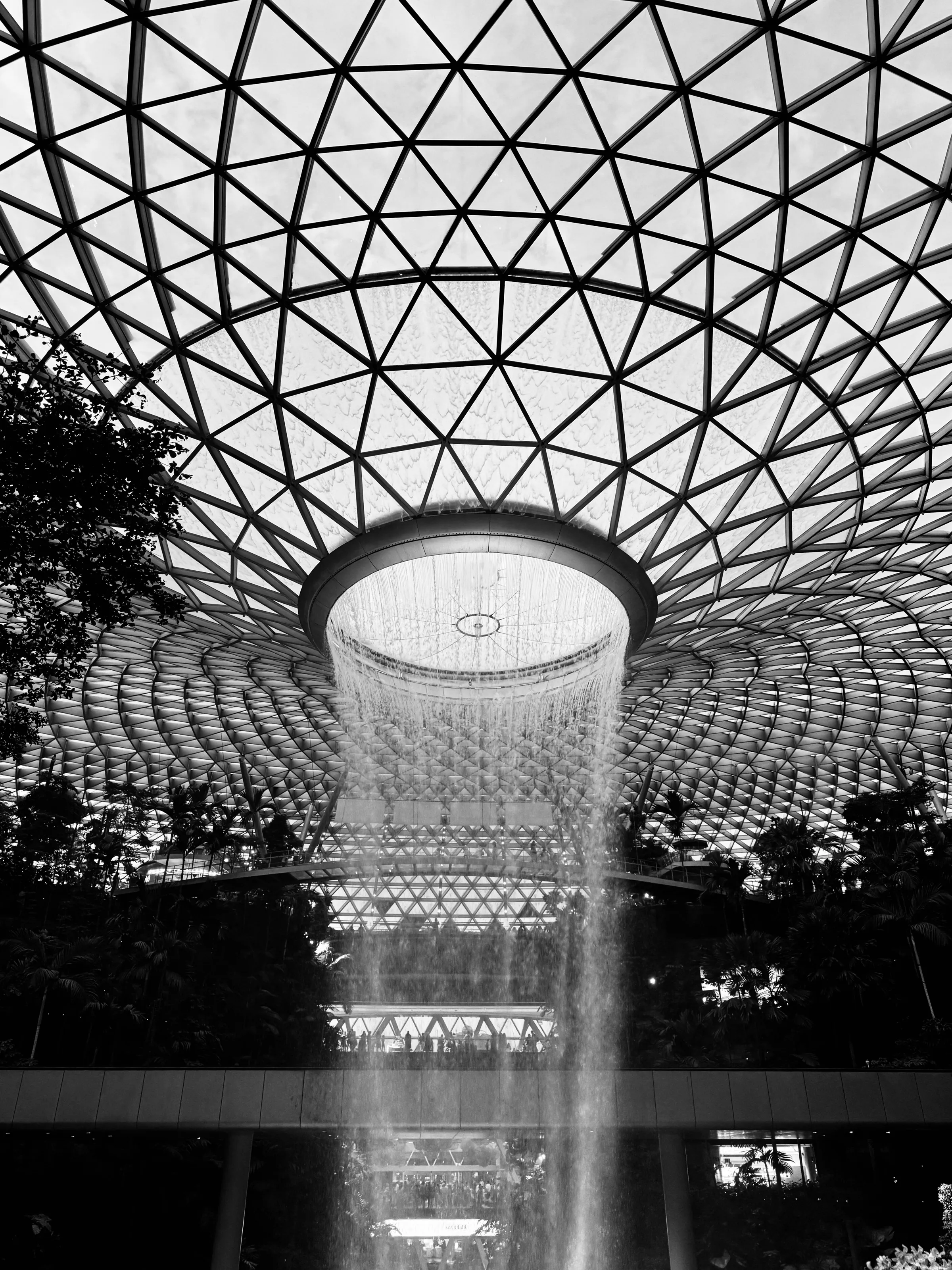 A tall indoor waterfall descends from a circular opening beneath a geometric glass roof.