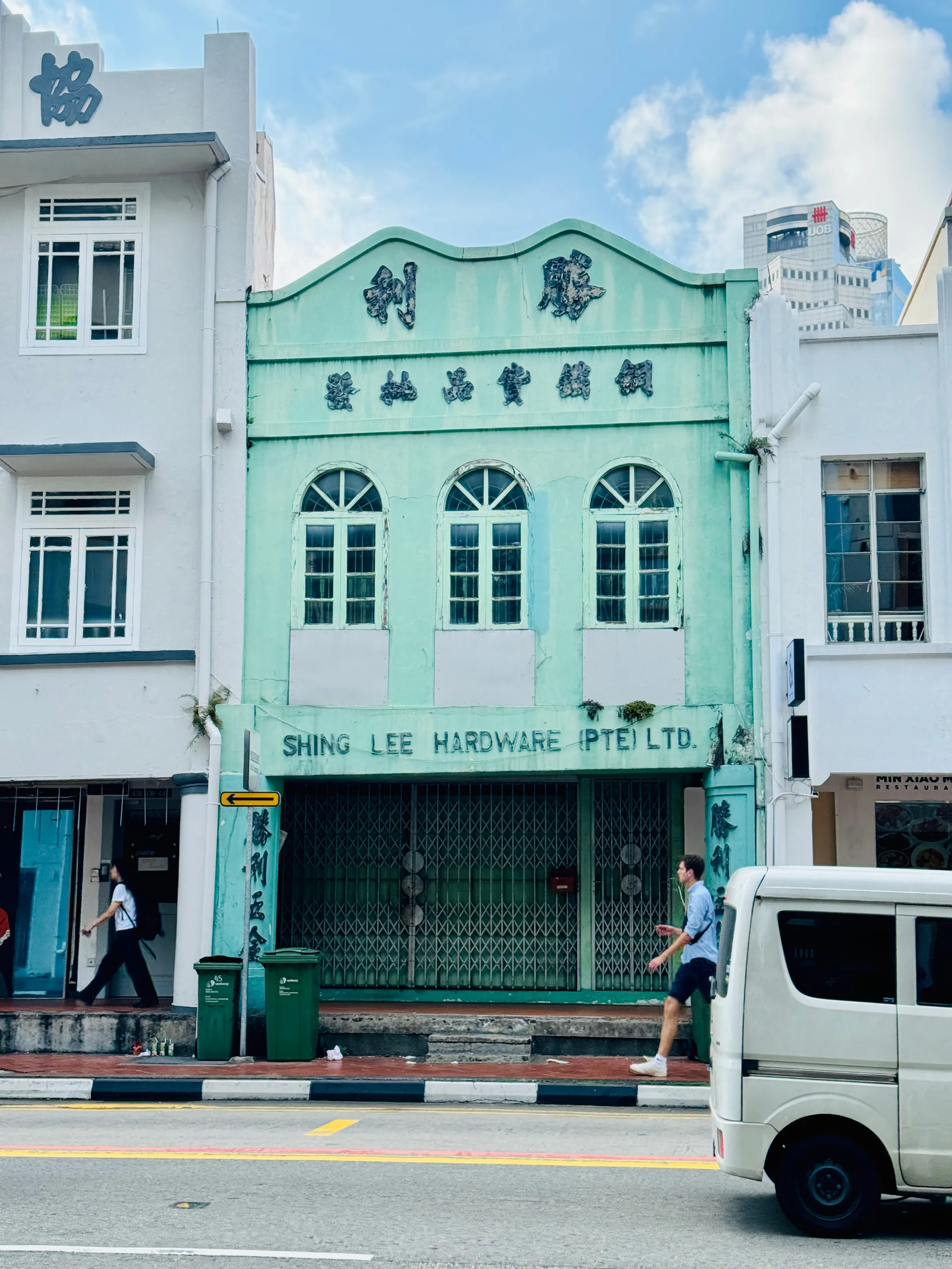 Street scene with a mint-green building labeled Shing Lee Hardware, two pedestrians, and a white van in the foreground.