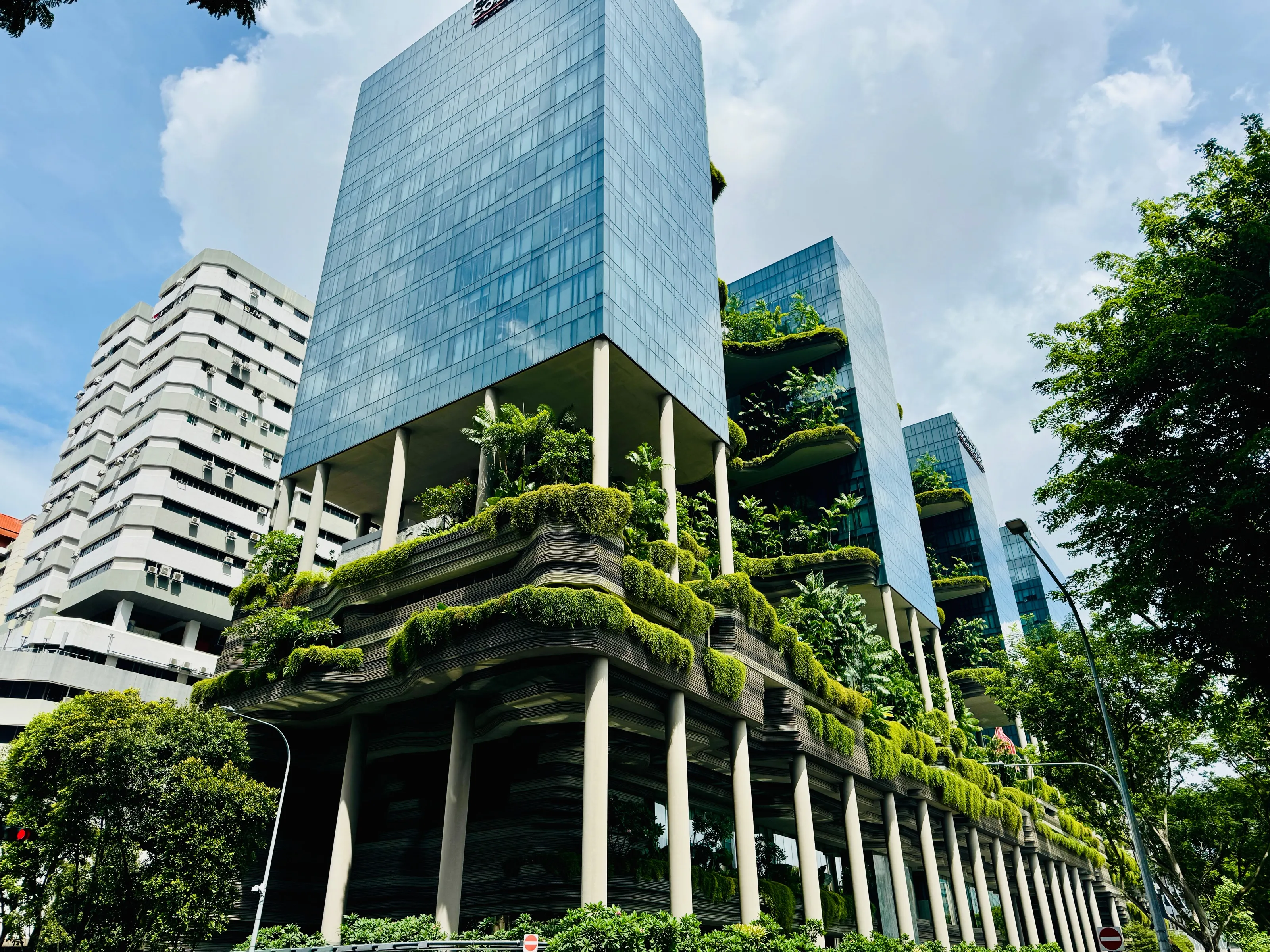 Low-angle view of a modern glass building raised on white columns above layered terraces covered in green plants.