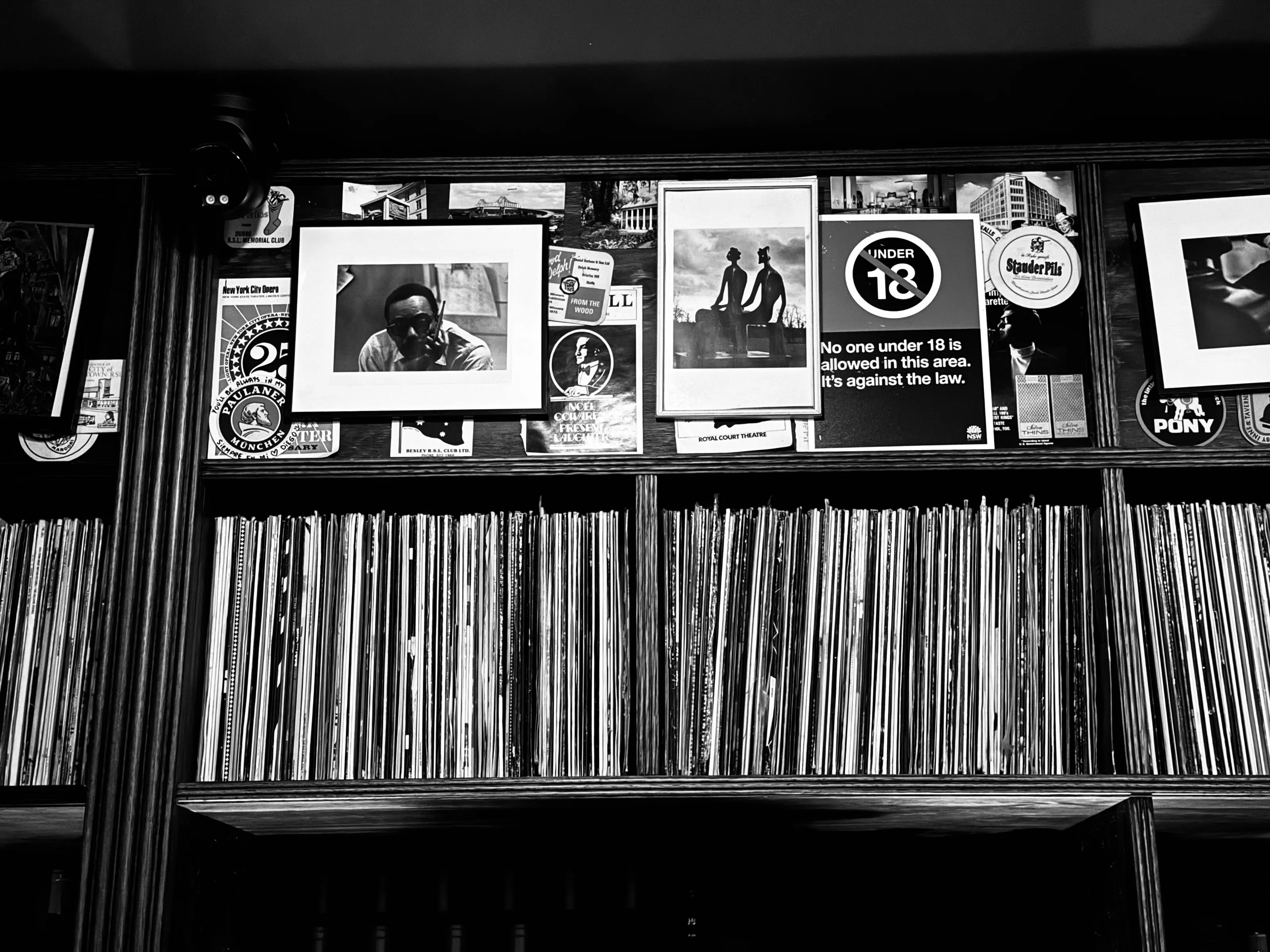 Black-and-white photograph of a wall covered with posters and signs above tightly packed vinyl records on shelves.