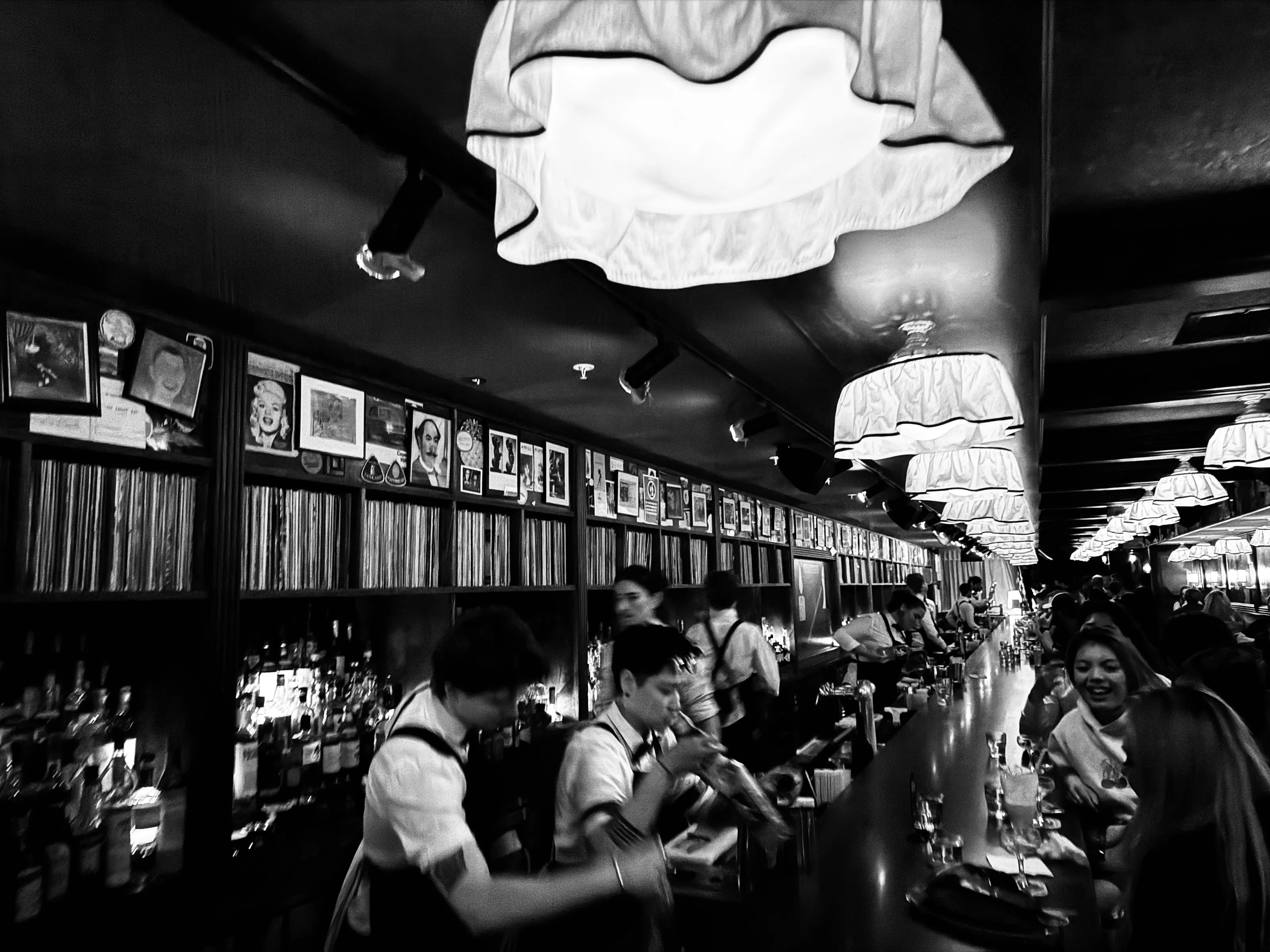 Black-and-white view down a long bar with bartenders at work, seated patrons, and hanging lanterns.