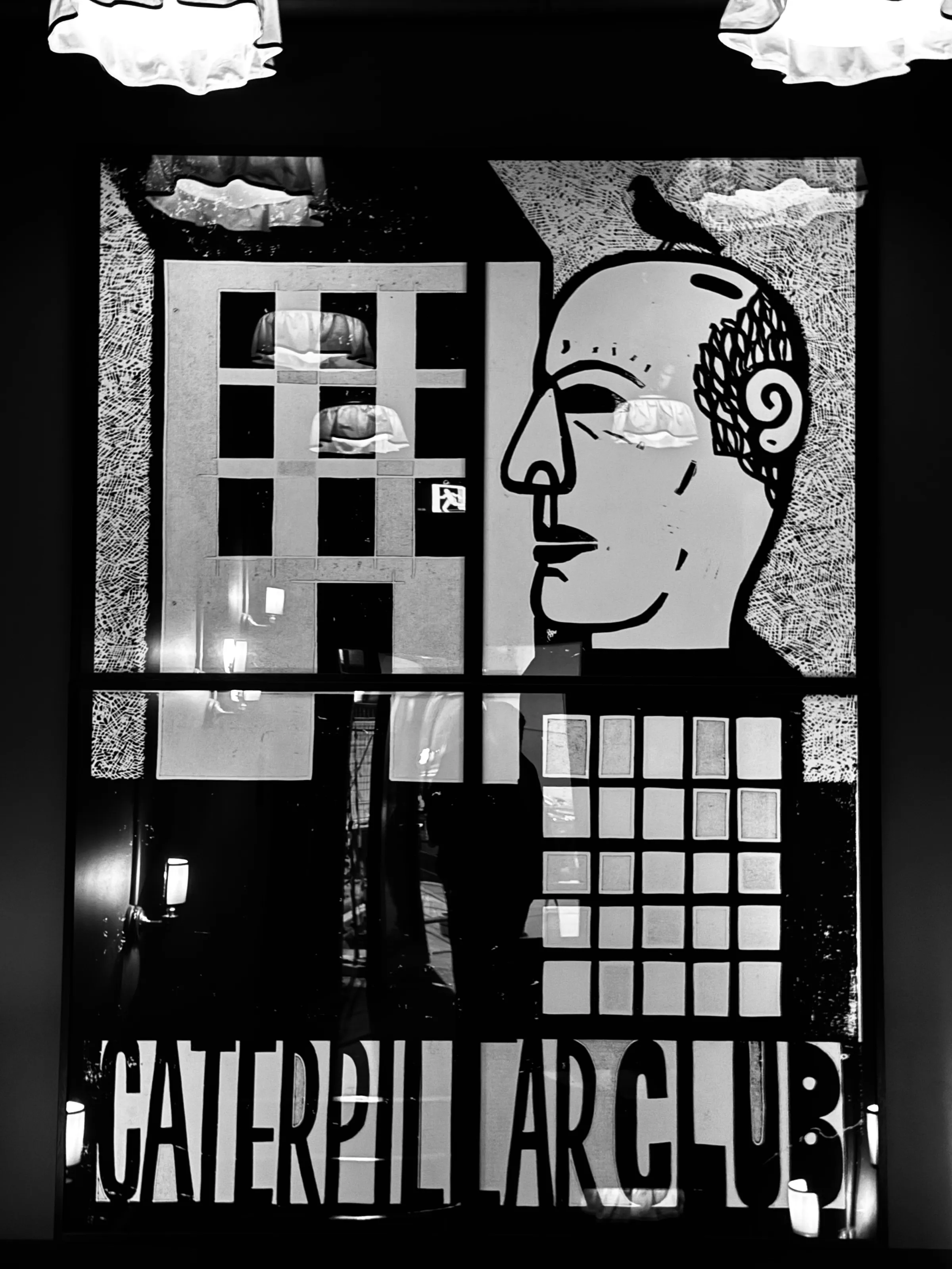 A black-and-white window display shows a stylized face, geometric shapes, reflected lights, and the words “Caterpillar Art Club.”