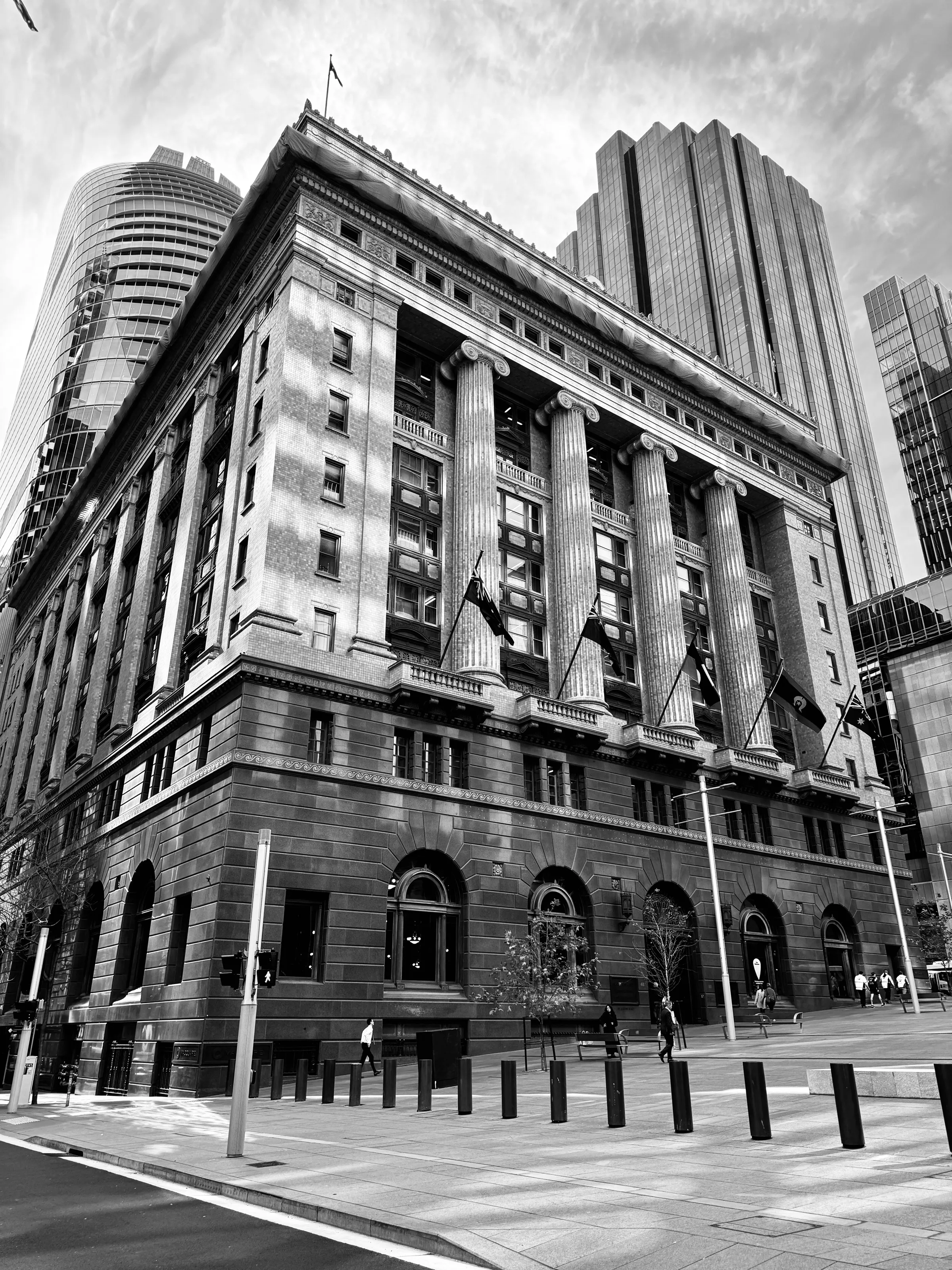 A large classical building with tall columns and flags stands on a city street amid high-rise towers.