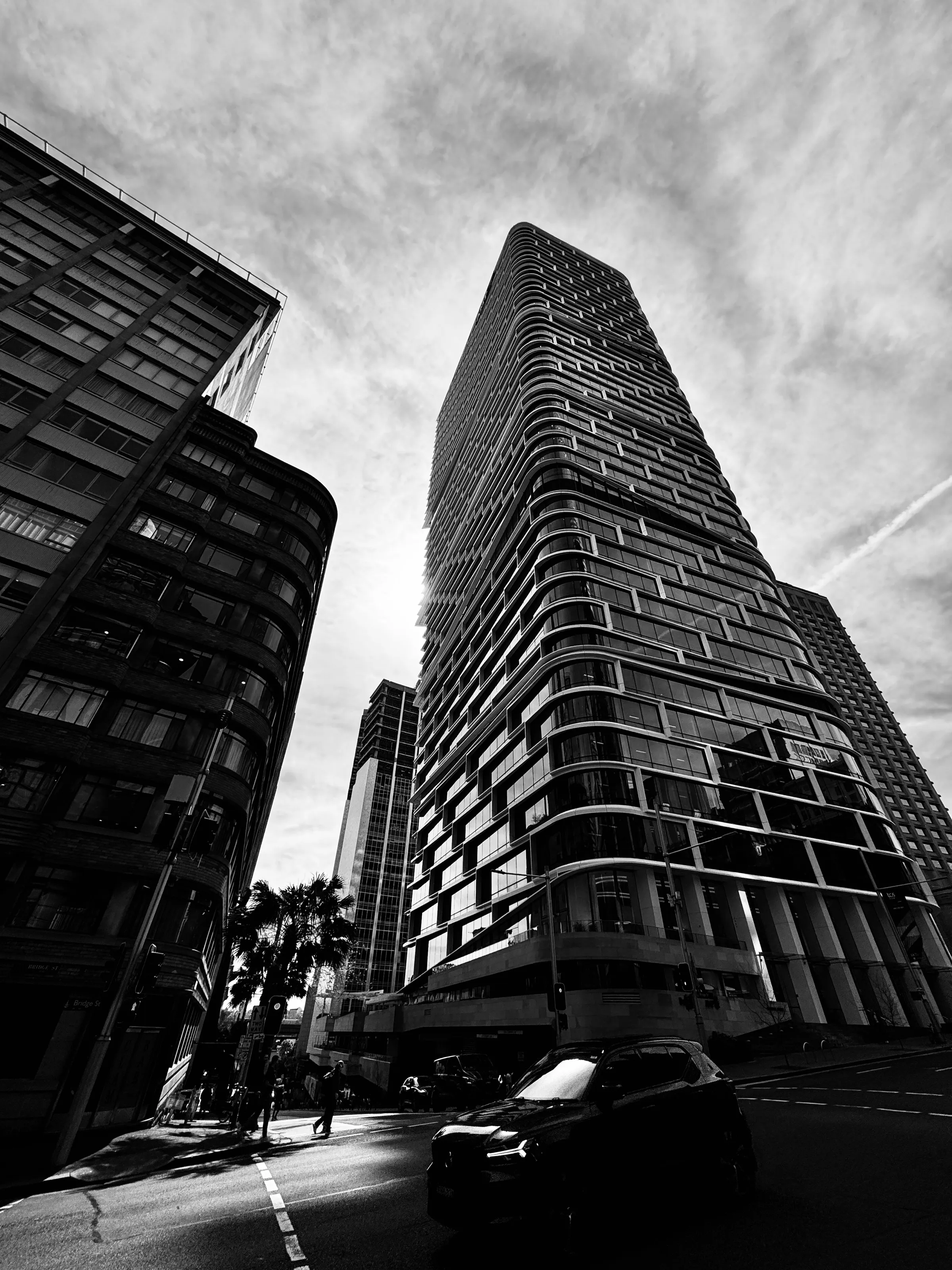 A tall rounded apartment tower rises between older buildings above a street with a car and pedestrians.