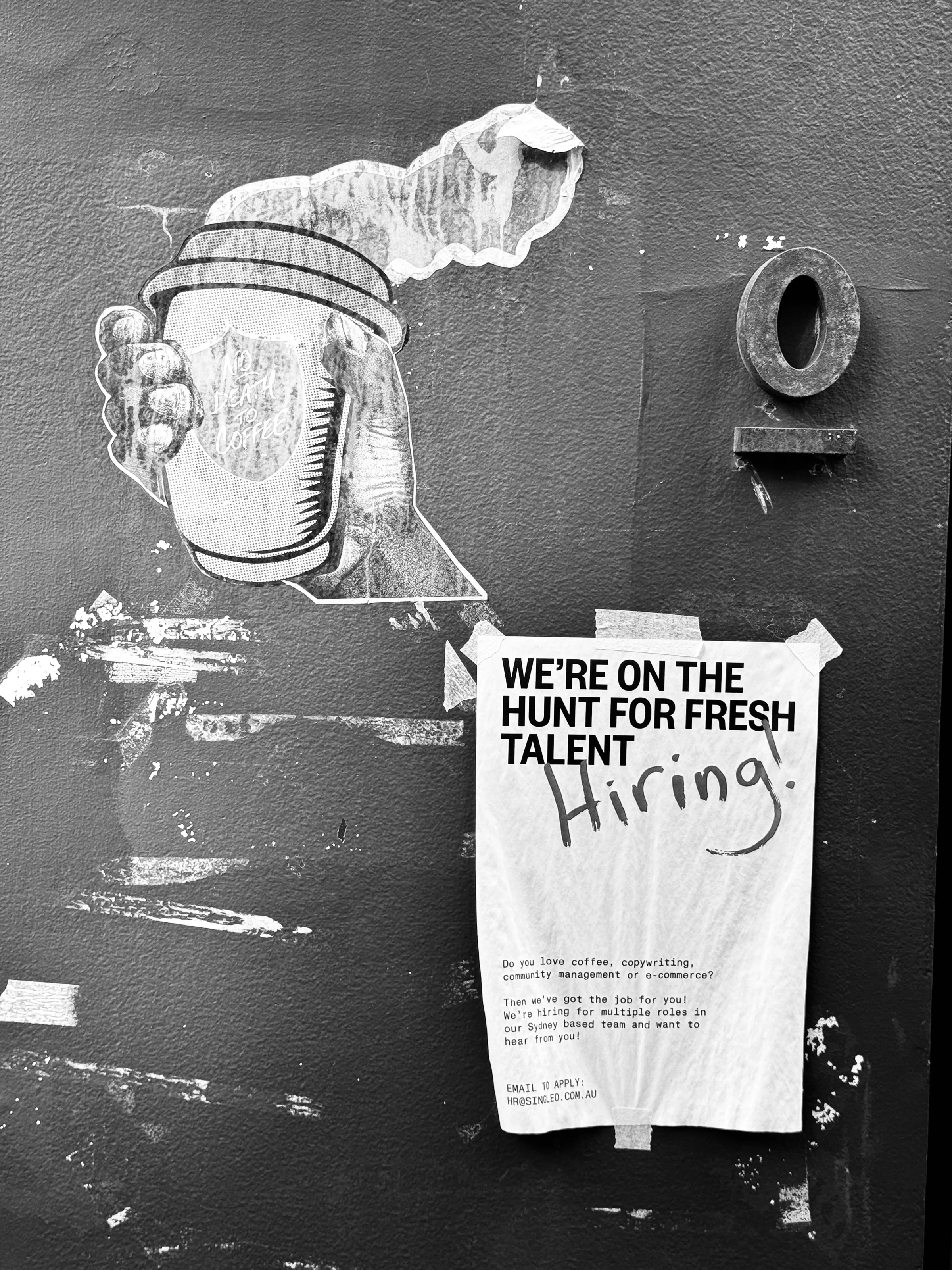 Black-and-white photograph of a wall with a painted hand holding a jar, a round opening, and a taped hiring notice.