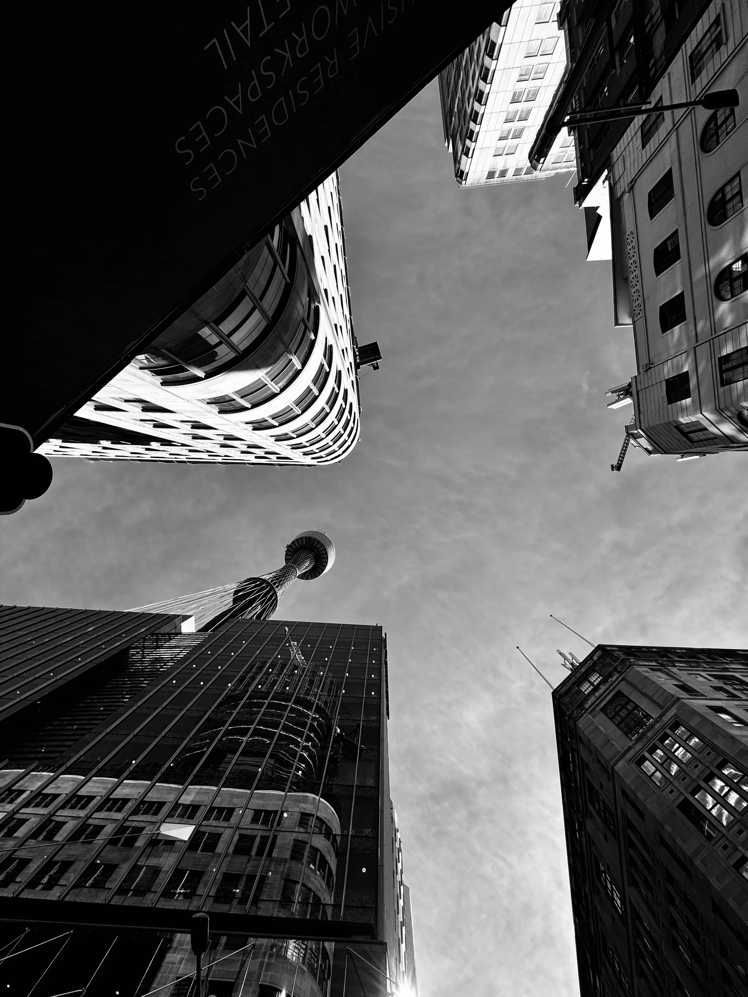 A black-and-white upward view between several tall city buildings, with a tower visible near the center.