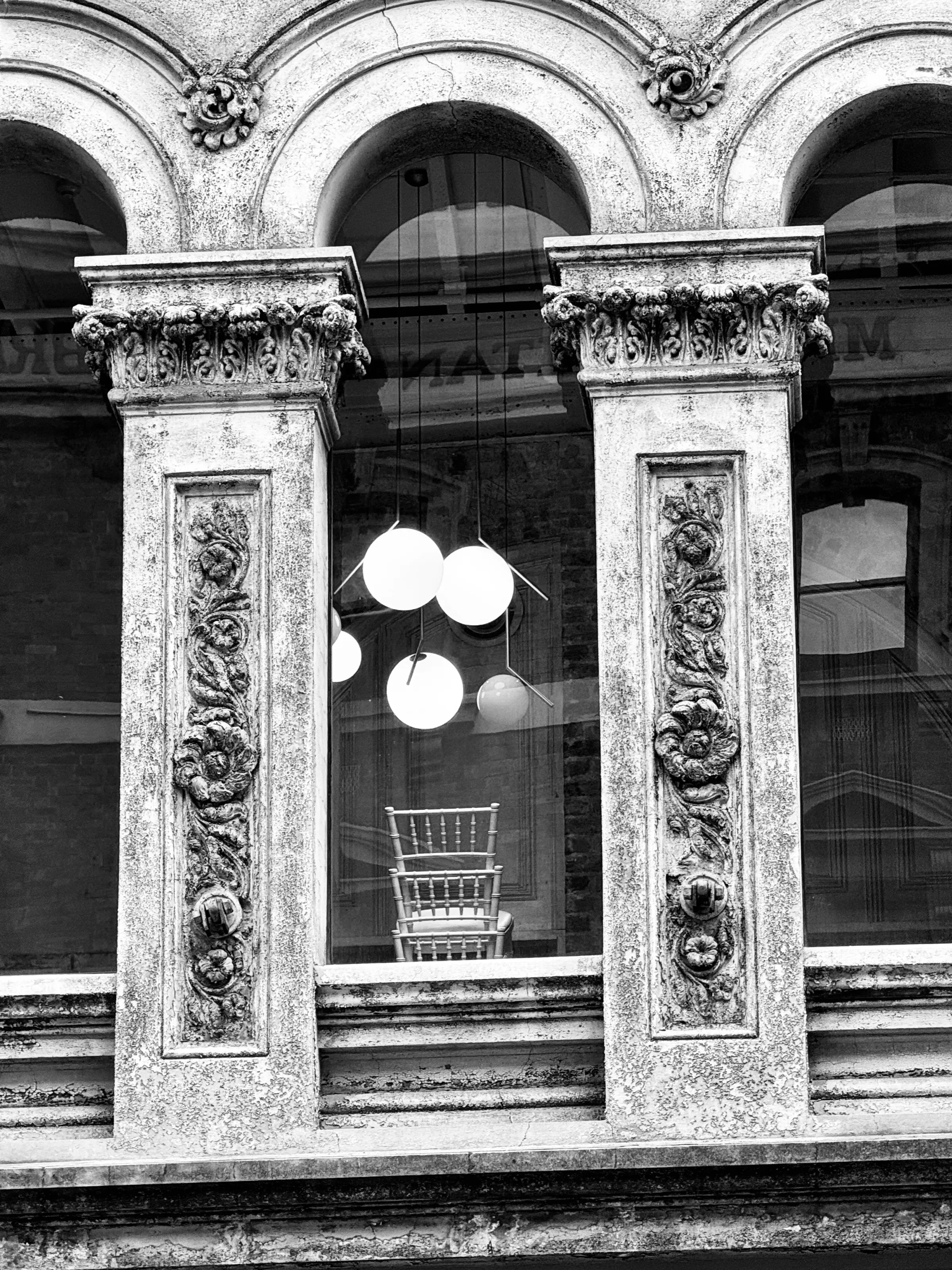 Black-and-white photograph of ornate stone columns framing a window with an empty chair and round hanging lights inside.