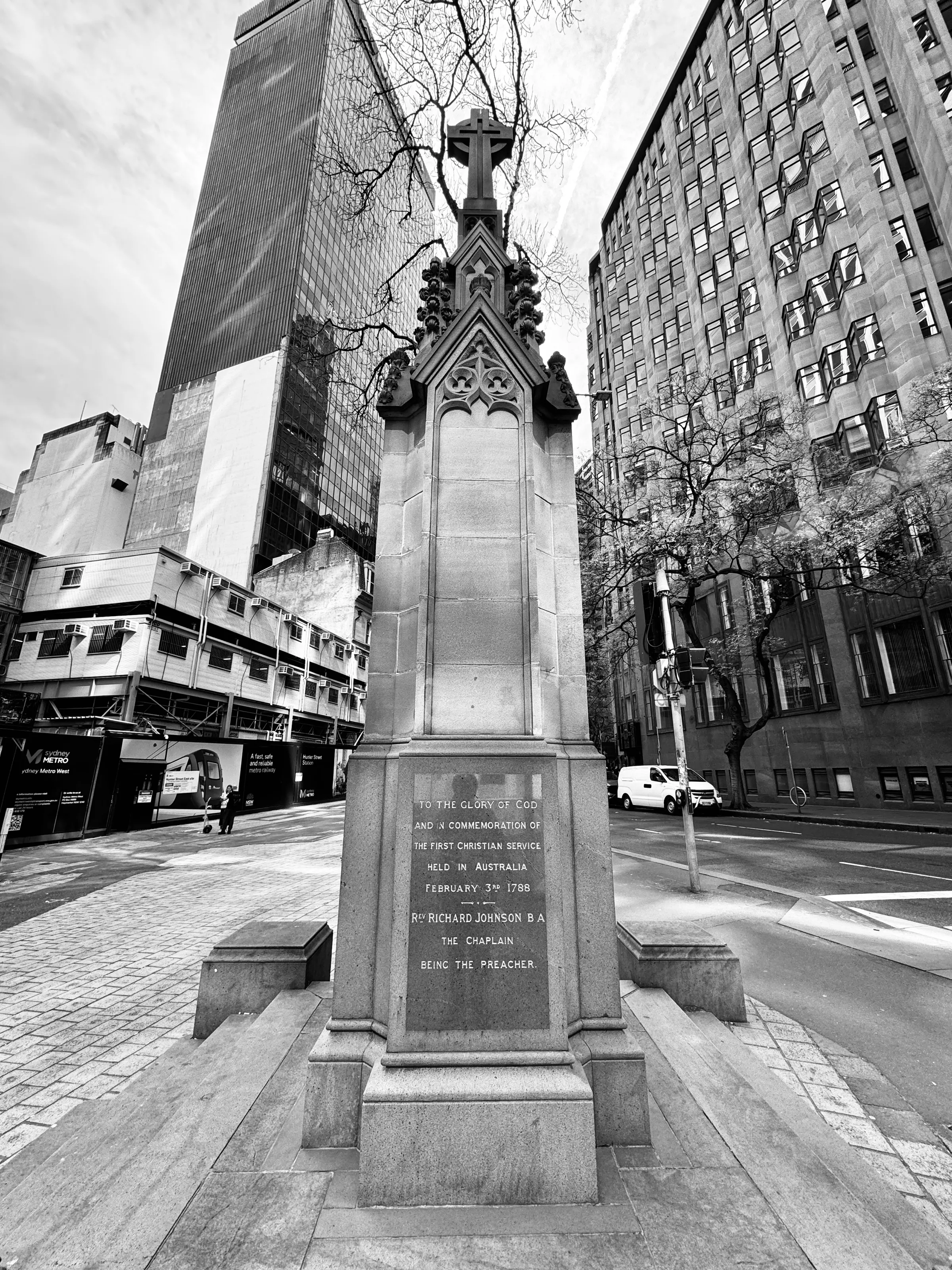 A Gothic stone memorial topped with a cross stands in a city street between tall buildings.