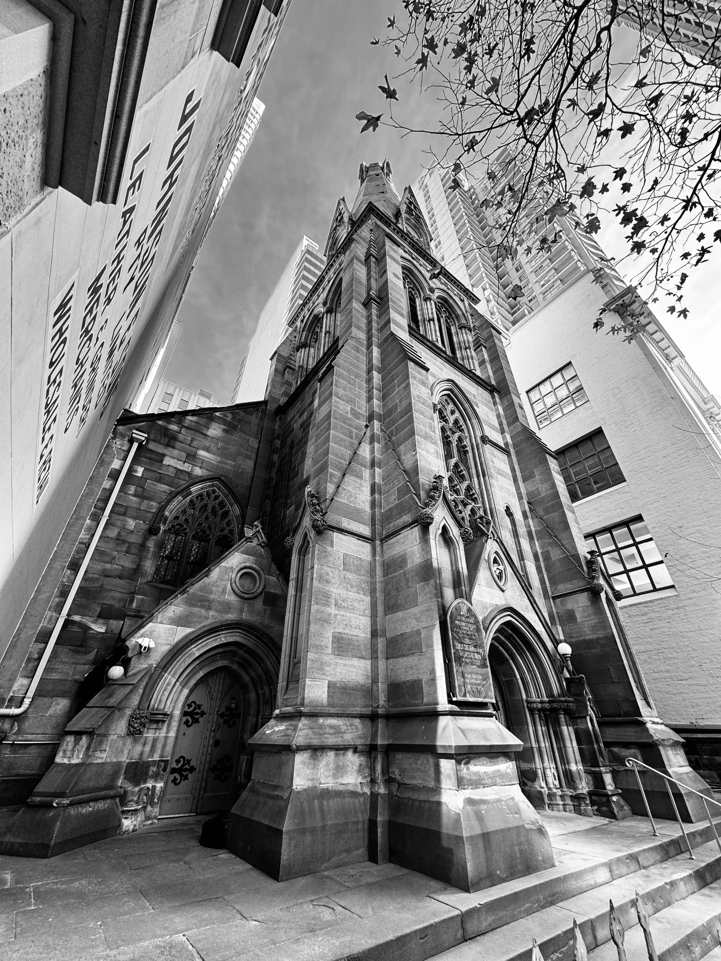 A stone Gothic church tower rises between modern city buildings, viewed from the sidewalk.