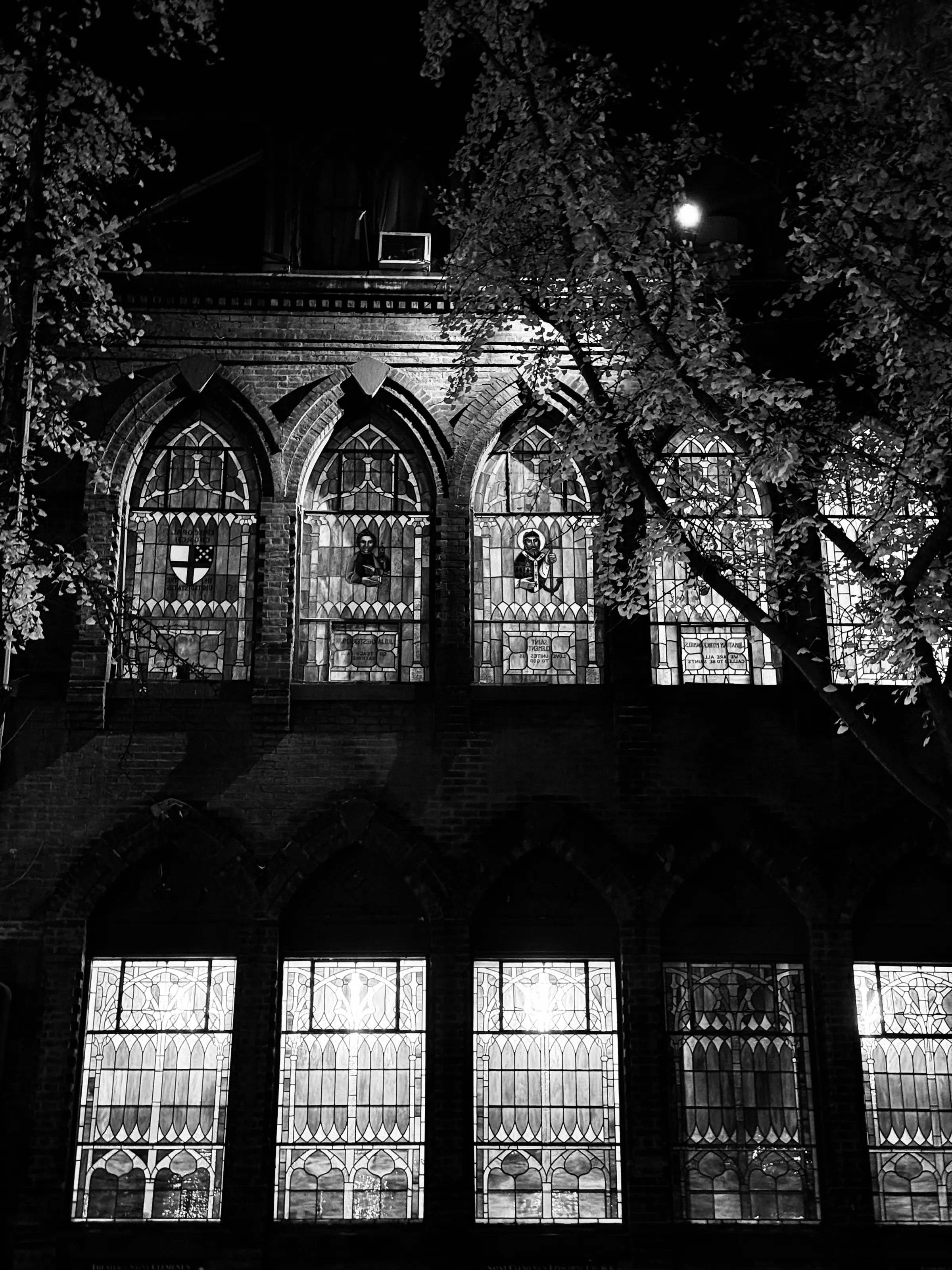 Black-and-white night photograph of tall stained-glass church windows partly obscured by leafy branches.