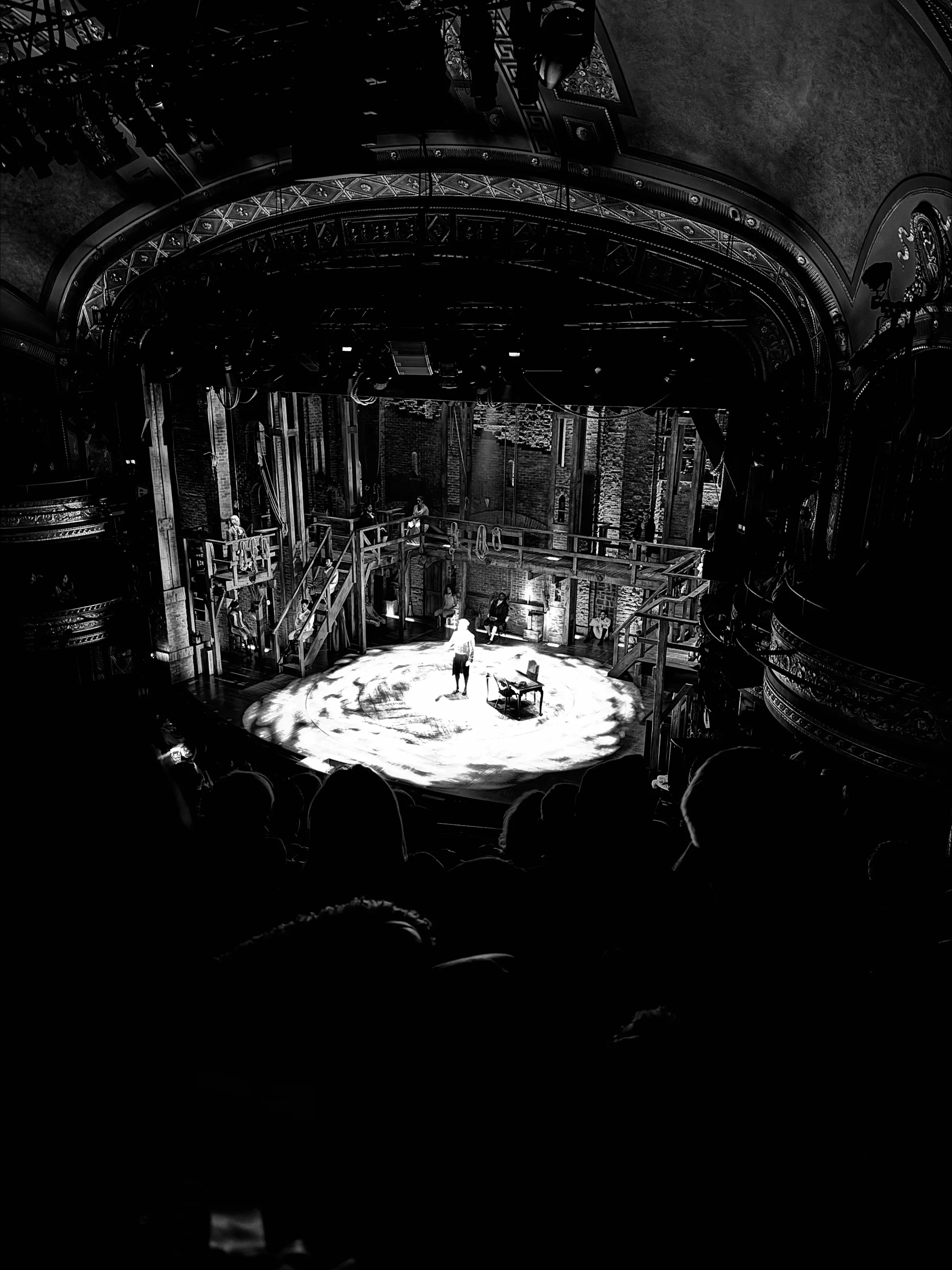 Black-and-white view from a theater balcony of a person in a bright spotlight on a stage, with audience silhouettes below.