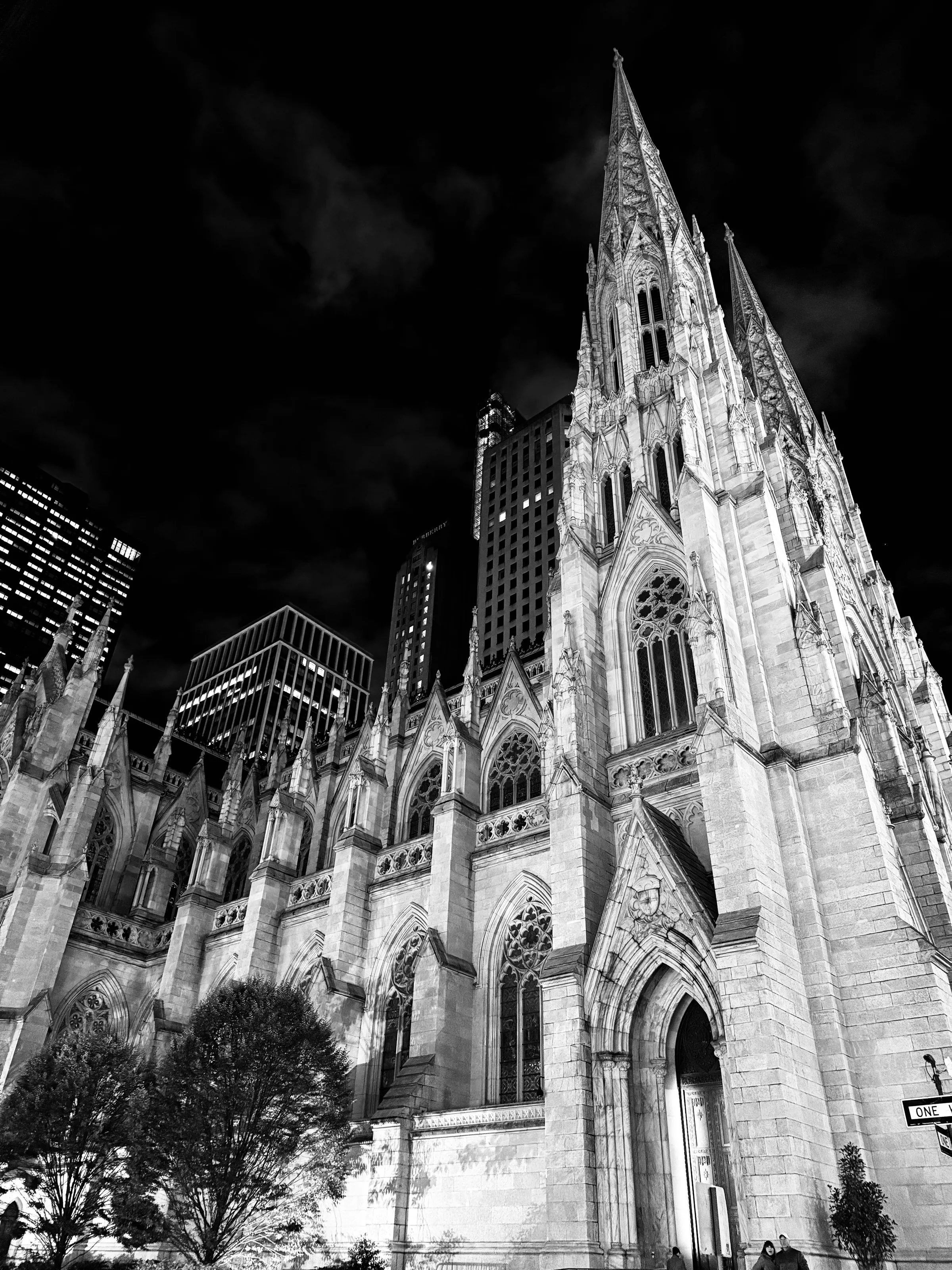 A large Gothic cathedral photographed from below at night, with modern buildings behind it.