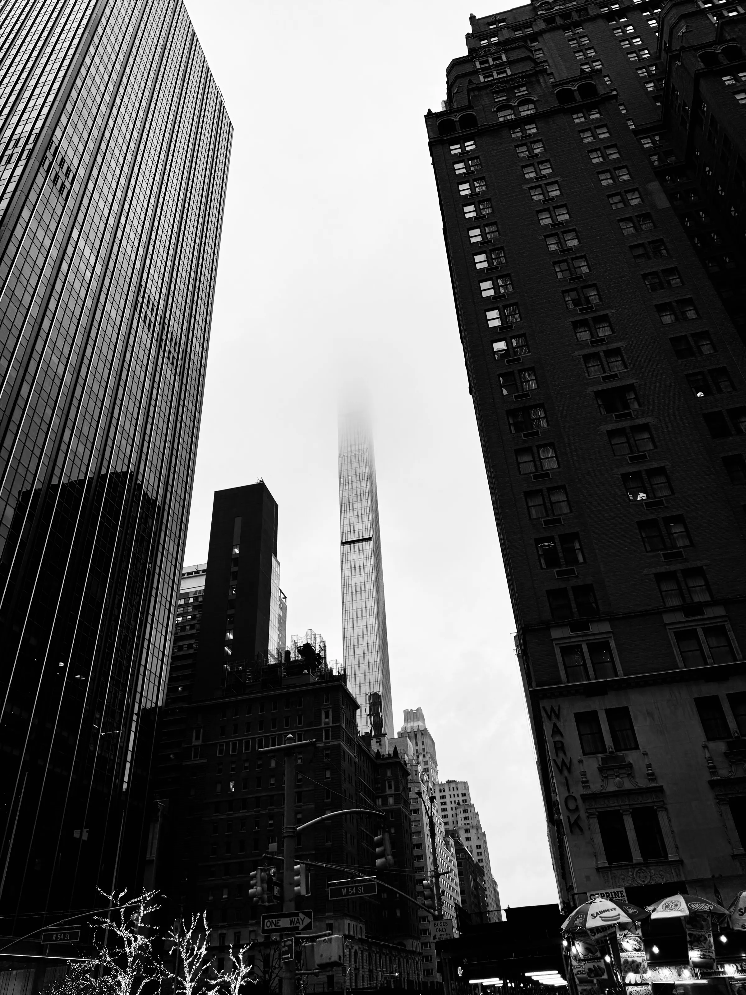 Black-and-white upward street view of tall buildings surrounding a slender tower whose upper section disappears into fog.