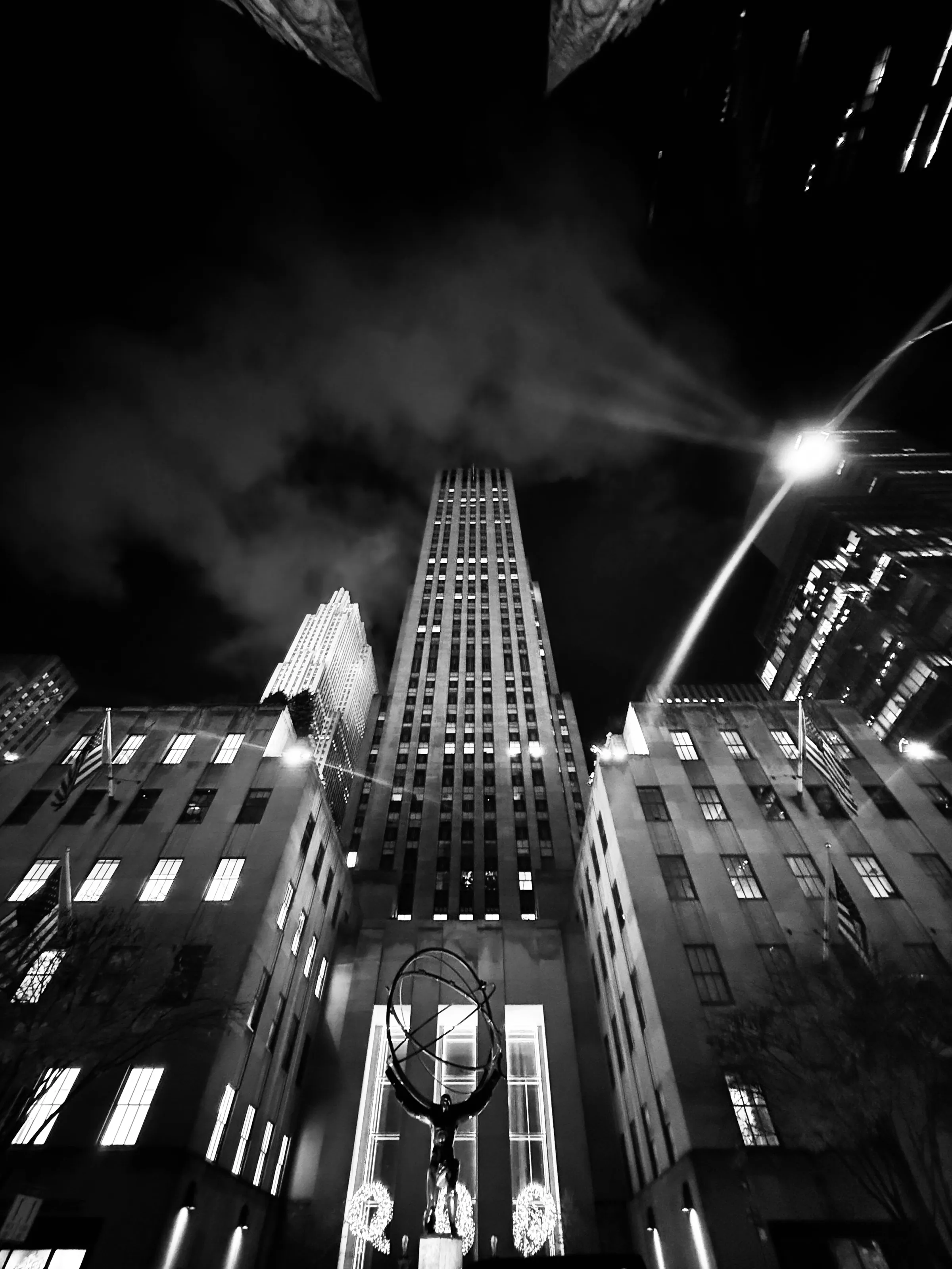 A tall illuminated building rises between lower office buildings at night in a black-and-white city view.