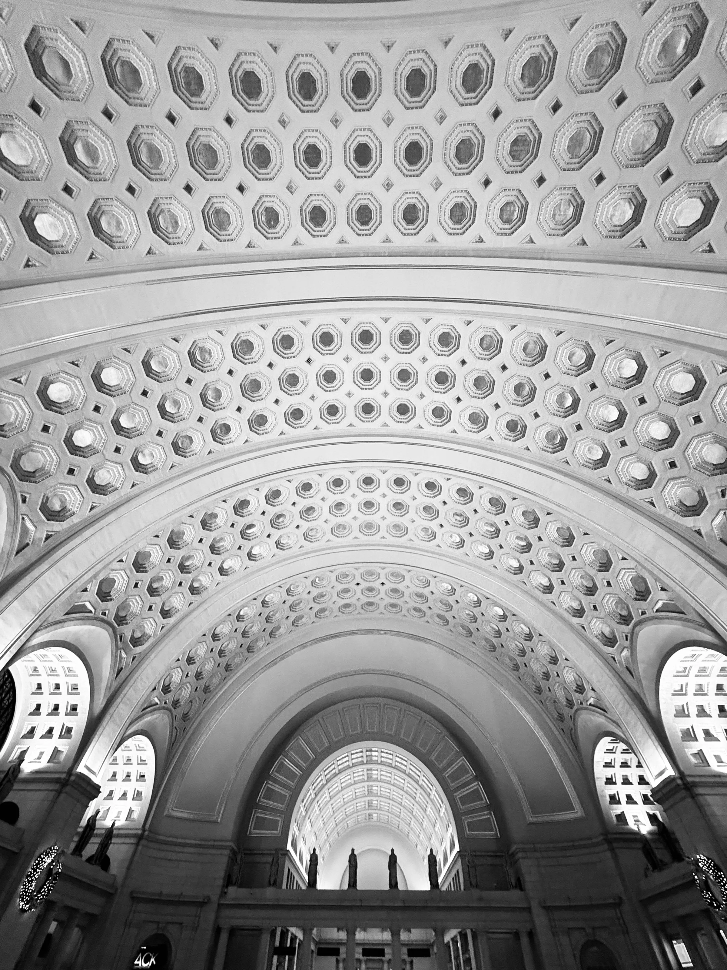 Black-and-white upward view through a grand hall with repeating coffered arches, patterned ceilings, and small silhouetted figures at the far end.