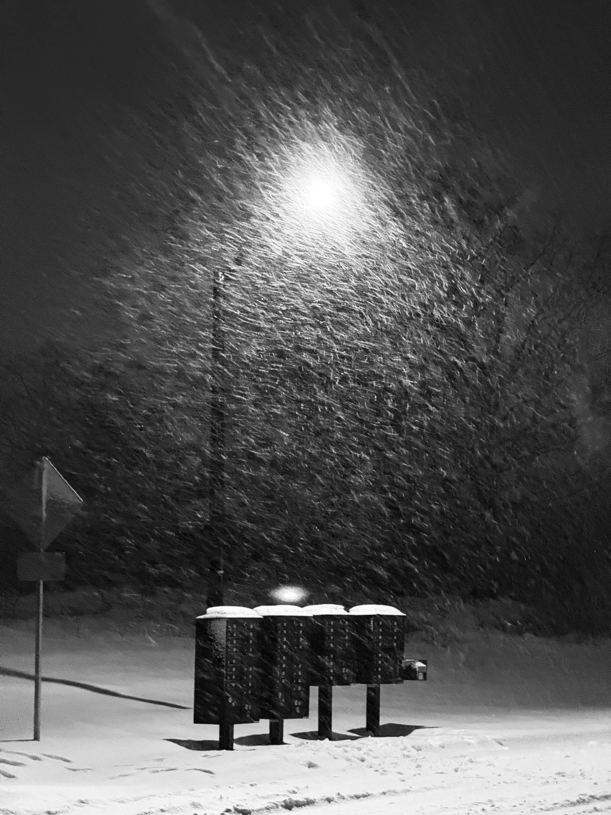 Black-and-white nighttime photograph of heavy snow blowing around a streetlight and three snow-covered mailboxes.