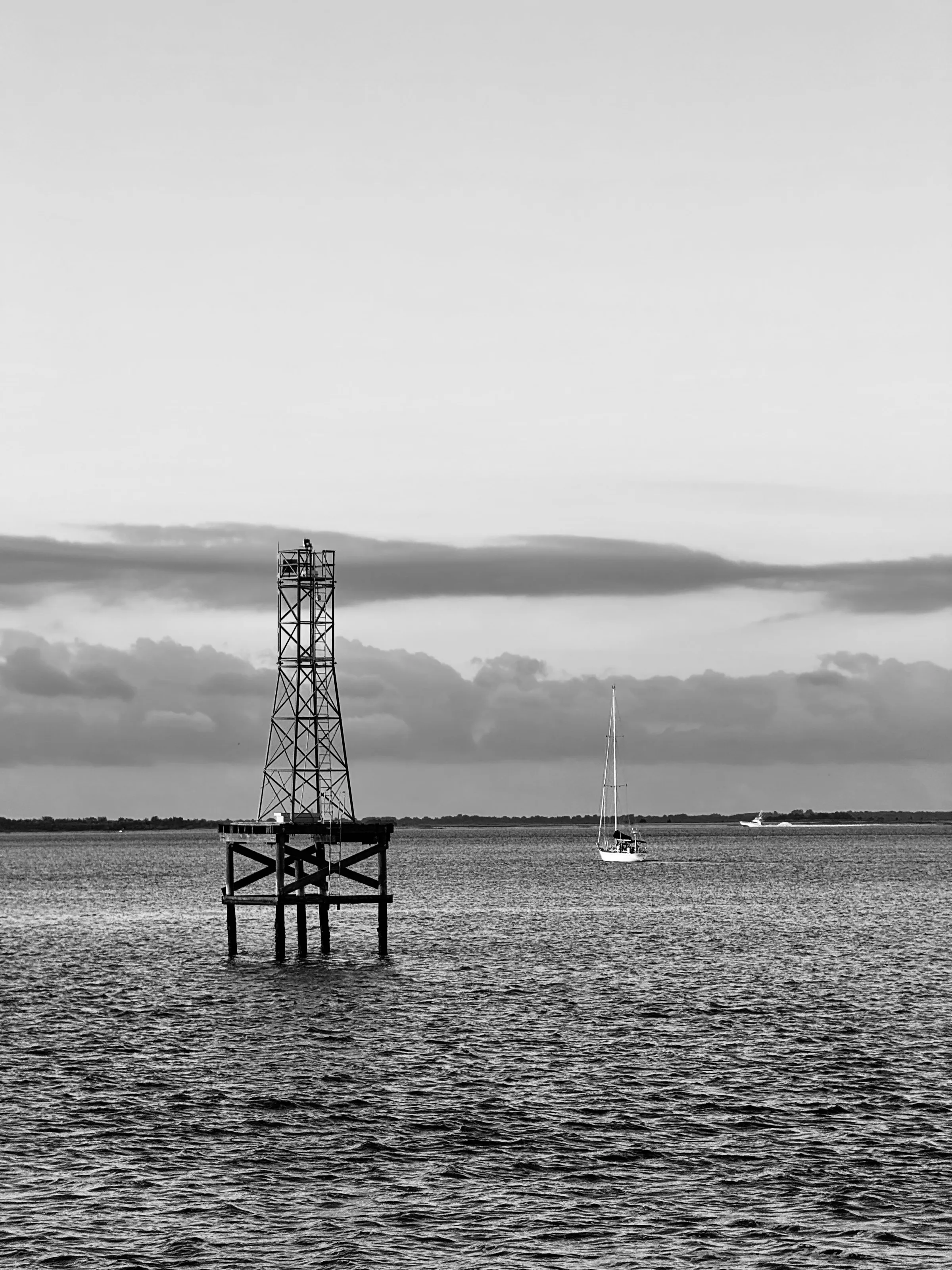 Black-and-white vertical photograph of a metal navigation marker in water, with a small sailboat near the horizon.