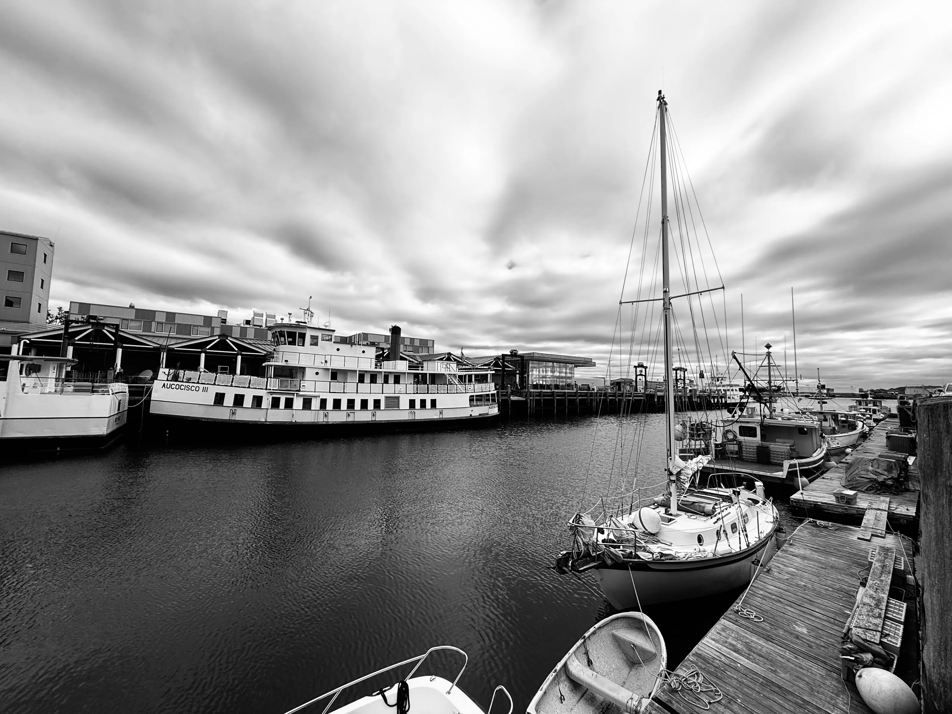 Several boats are tied beside wooden docks in a harbor under a cloudy sky.