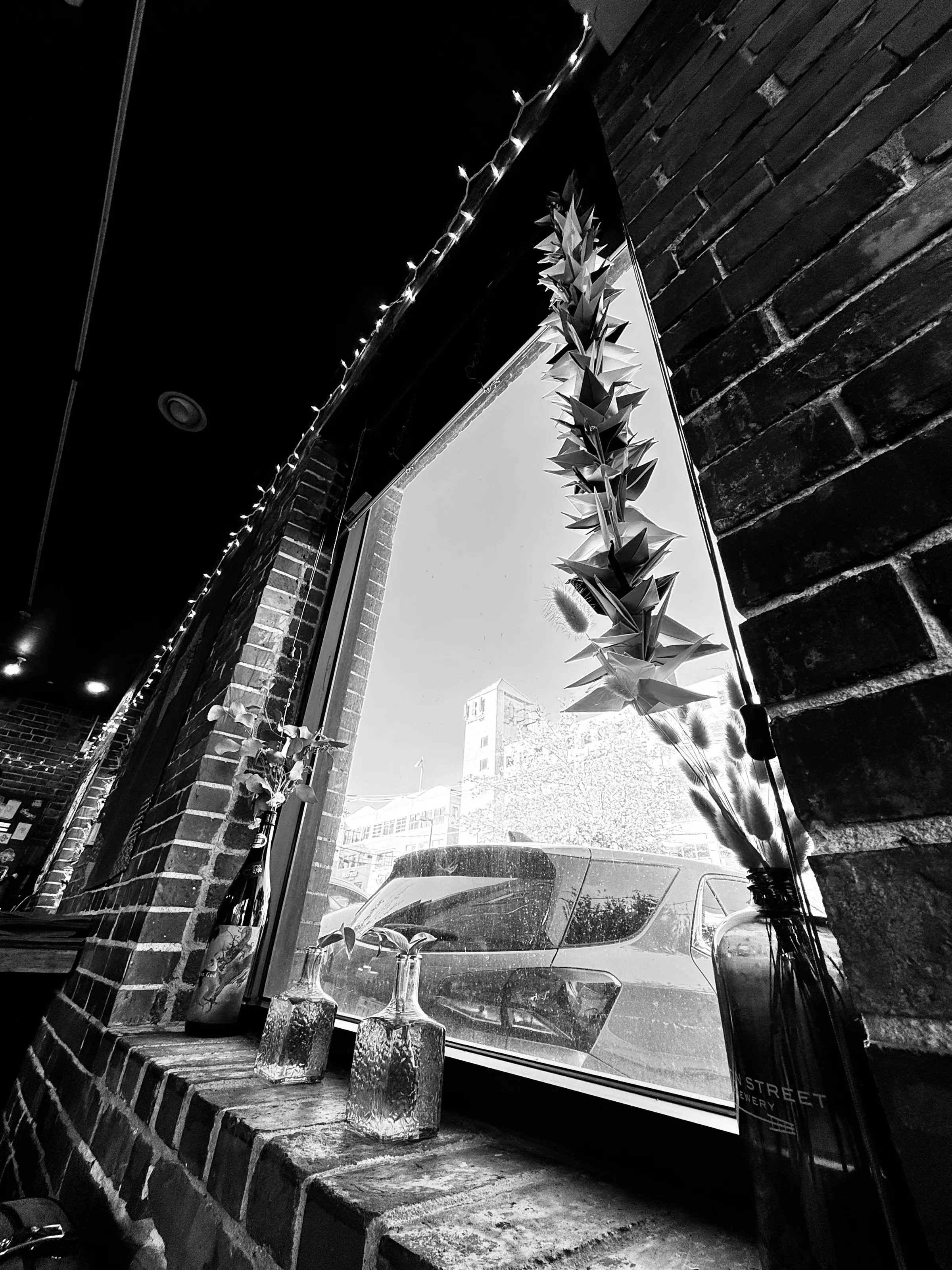 Black-and-white view from inside a brick building toward a bright window with hanging folded paper decorations, bottles, and a parked car outside.