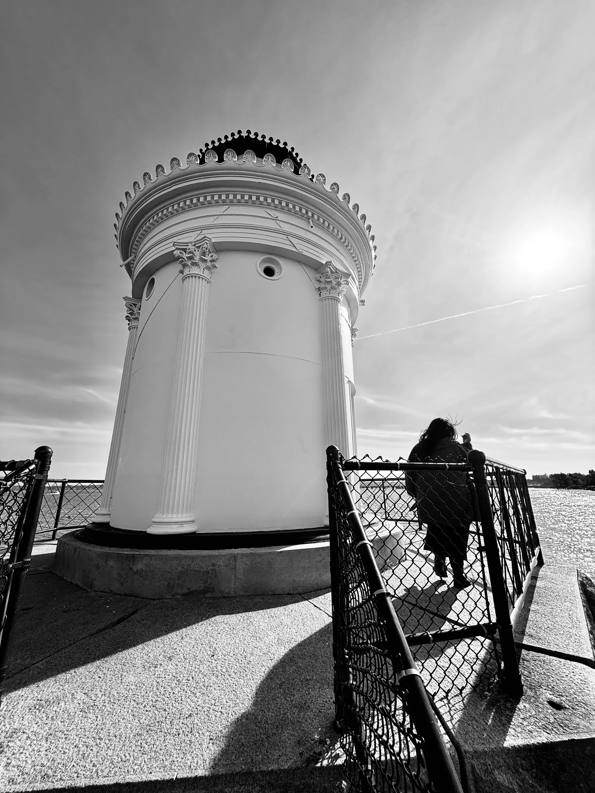 Black-and-white wide-angle photograph of a round white tower with columns beside water, with a person standing by a chain-link fence.