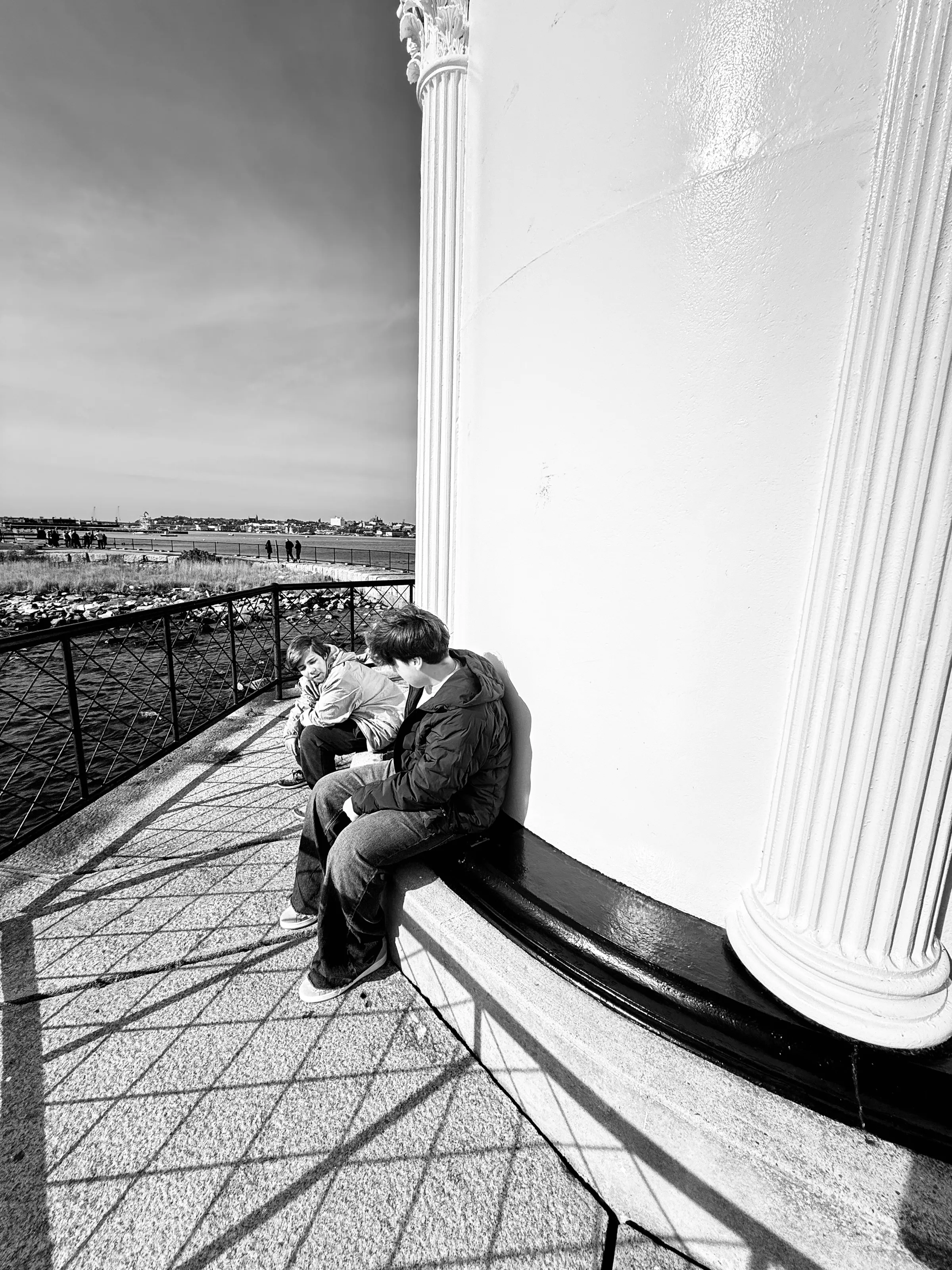 Black-and-white photograph of two young people sitting on a ledge beside a tall white column, with long railing shadows across the pavement.