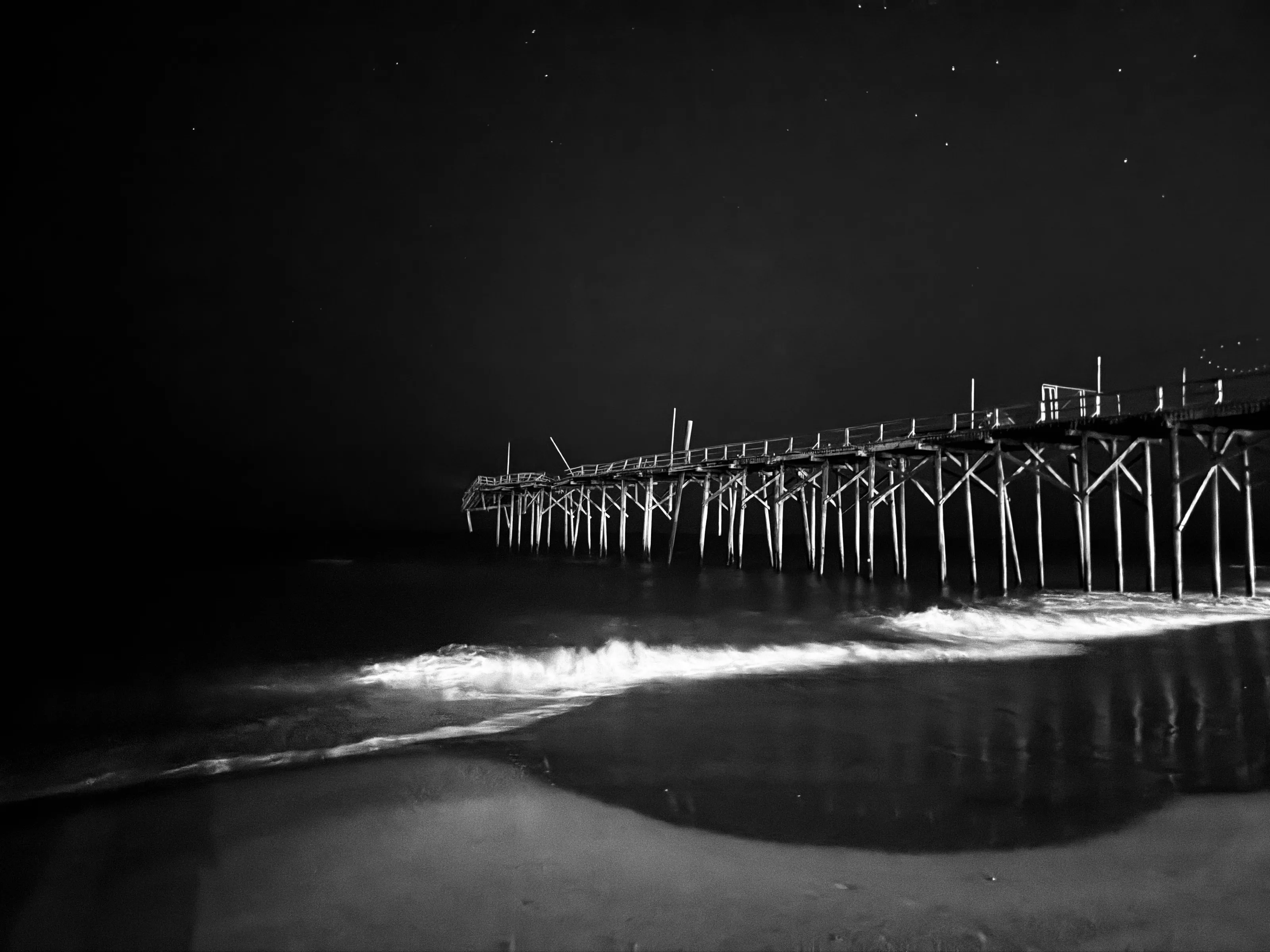 A black-and-white night view of a long wooden pier over dark waves, with a few stars overhead.